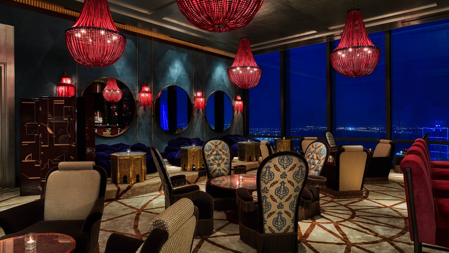 Cocktail tables in dimly-lit Blue Moon lounge under large red beaded chandeliers, high-back plush chairs with decorative prints