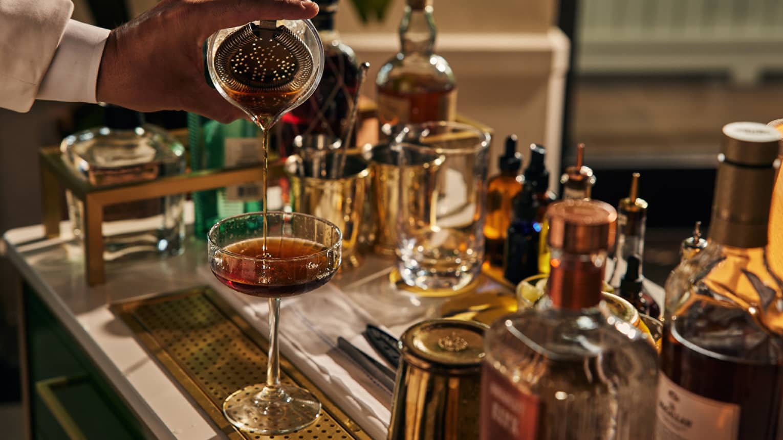 A bartender pouring a drink into a cocktail glass with bottles of liquor around.