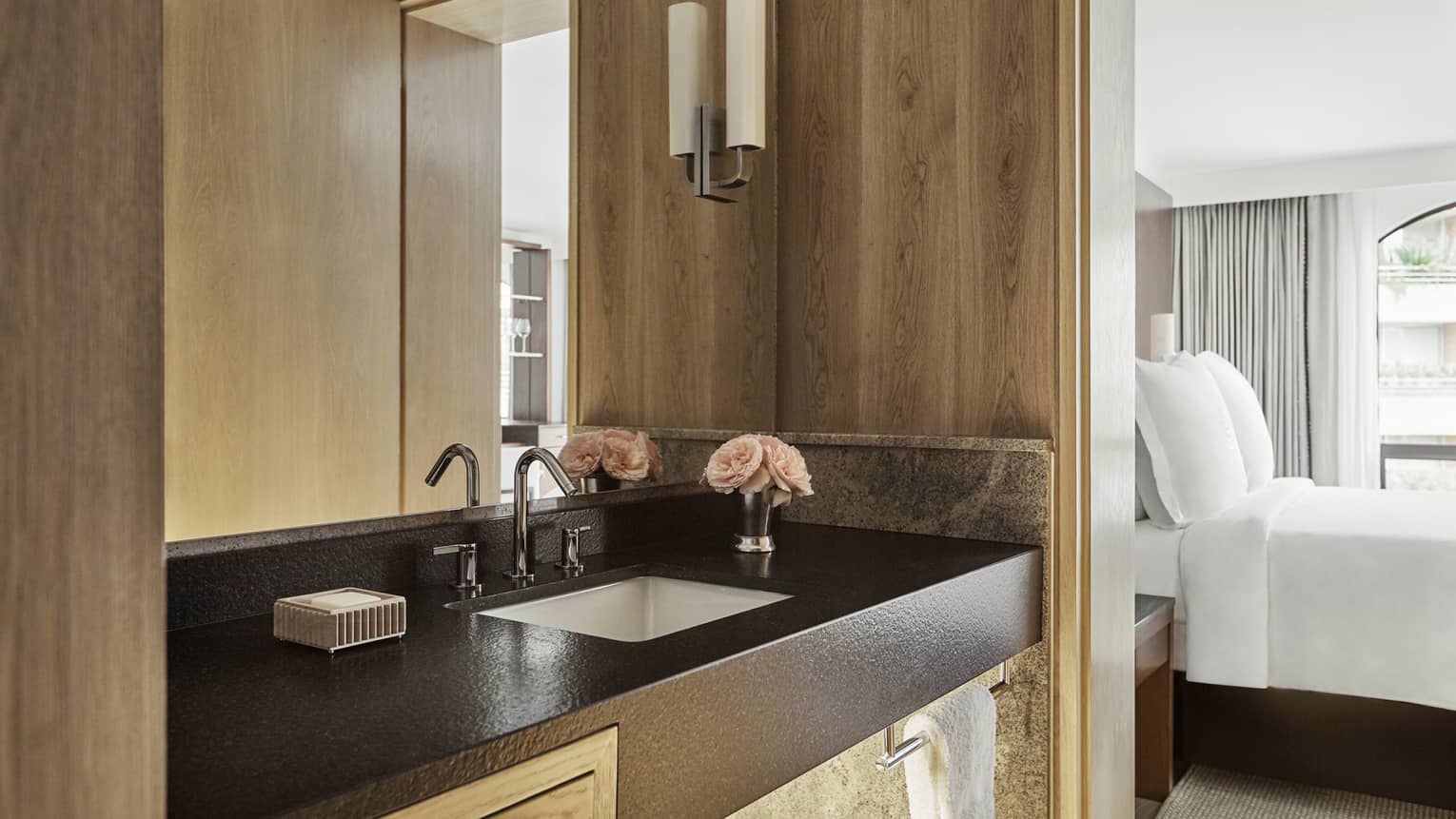 Close-up of bathroom vanity with black counter, sink, wood walls, bedroom visible to side