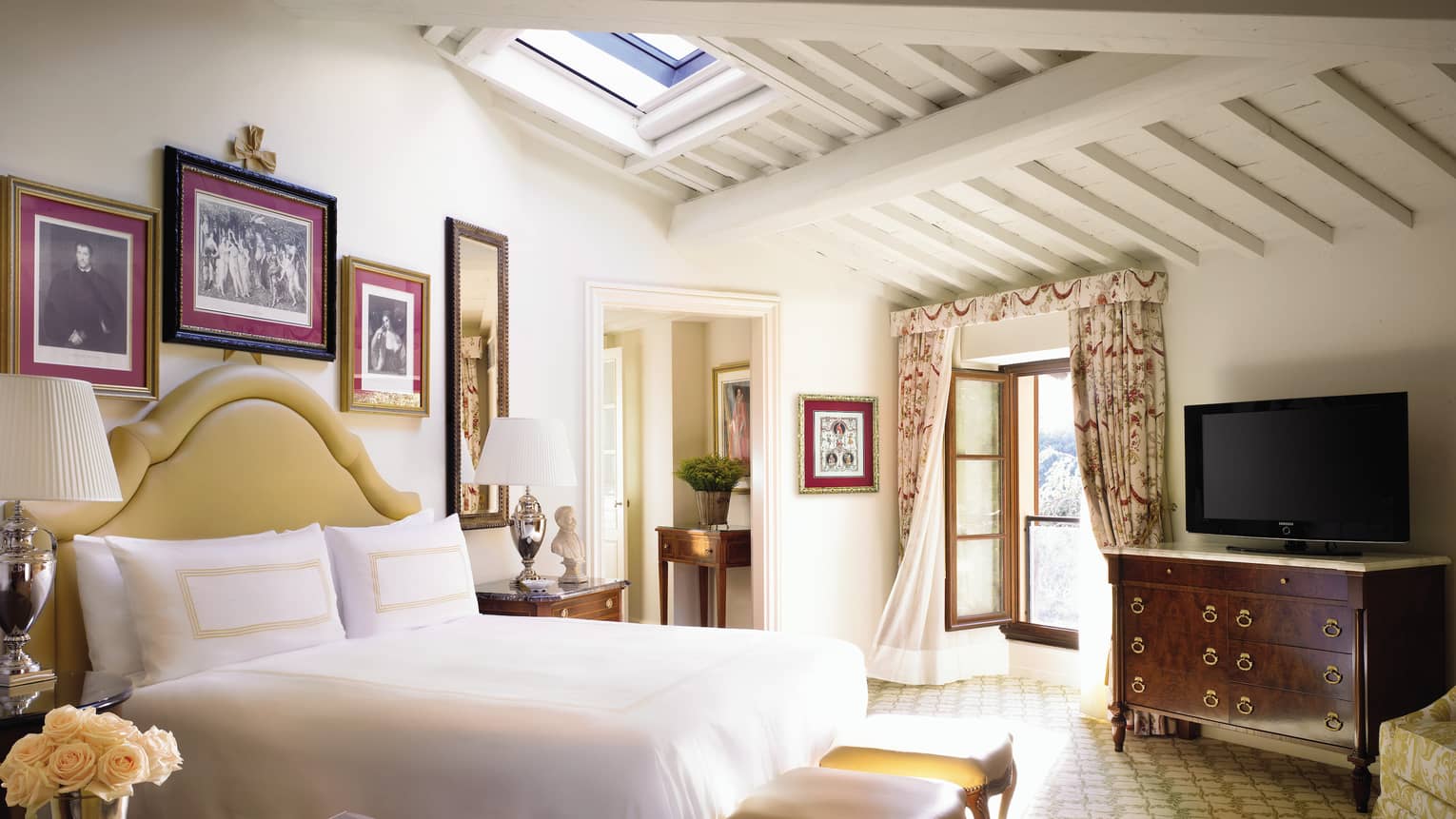 Hotel bed with padded headboard under sloped ceiling, skylight, open sunny window with flowing curtains