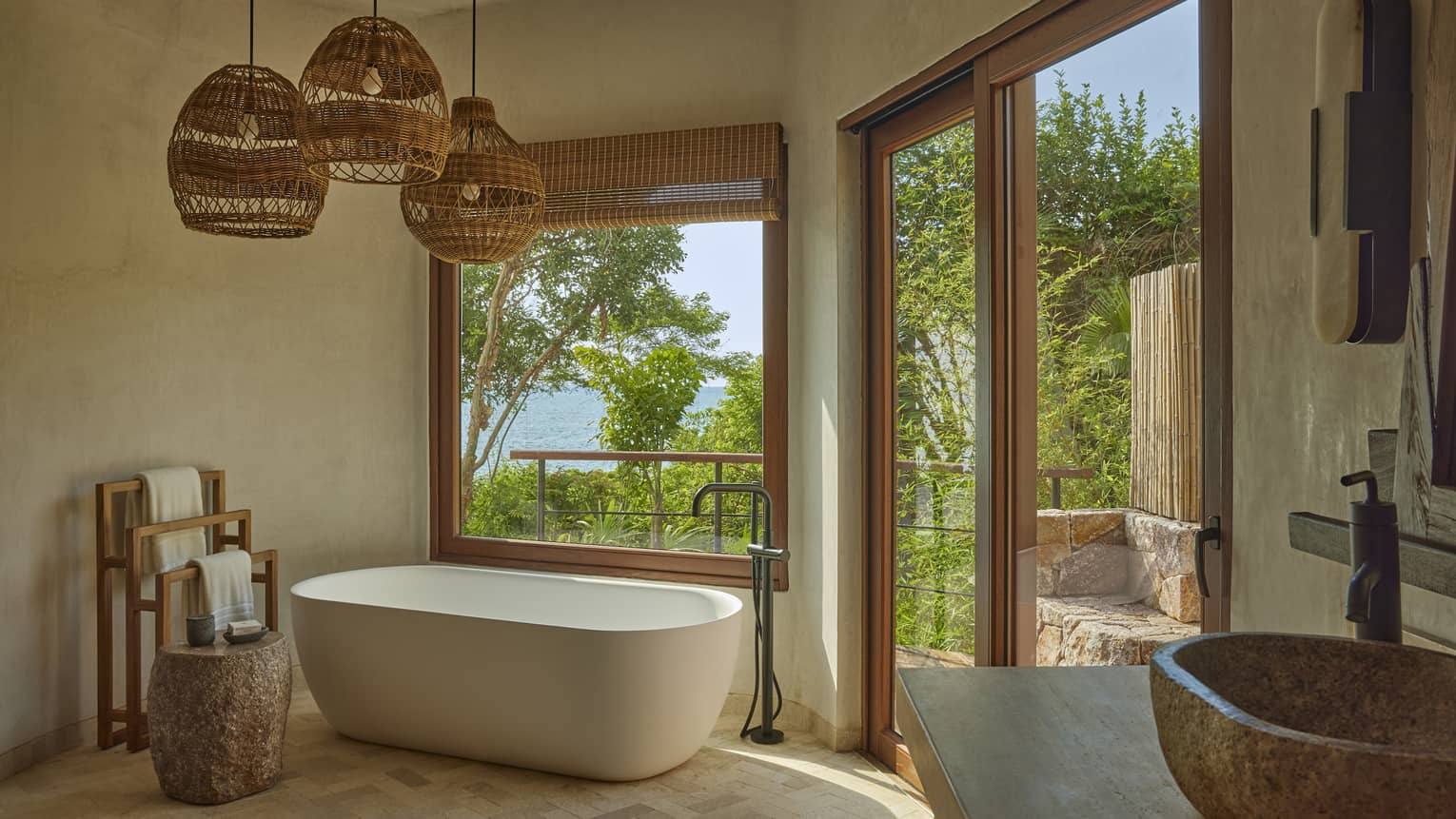 Bathroom with freestanding tub, three hanging whicker lights and window to tropical forest