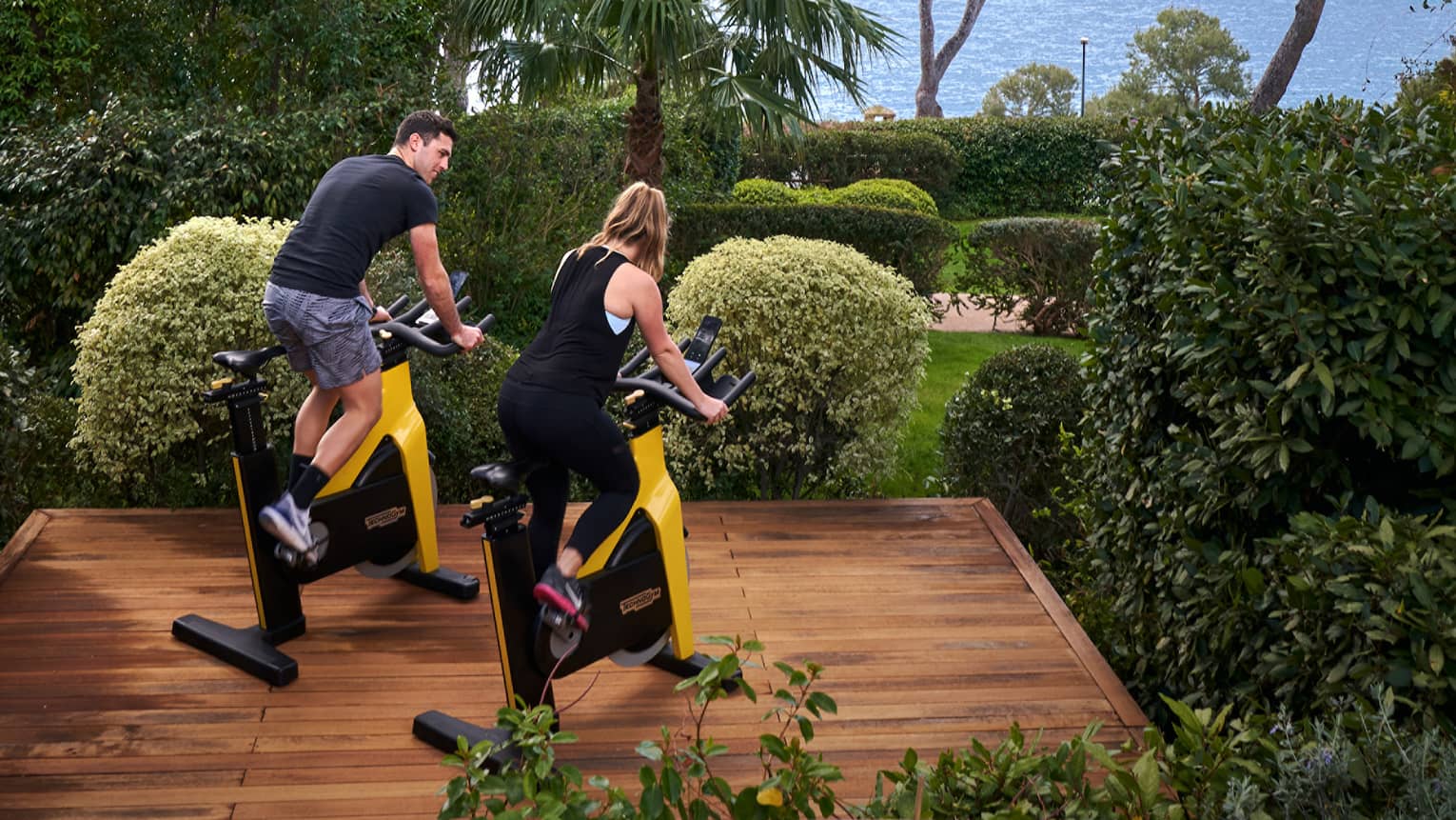 A man and a woman ride stationary bikes on outdoor wooden platform, sea in the distance