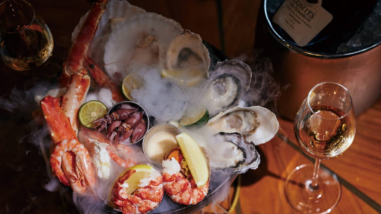 Seafood platter on wooden table with lobster, giant prawns, octopus, oysters and clams; glass of white wine and wine bucket nearby