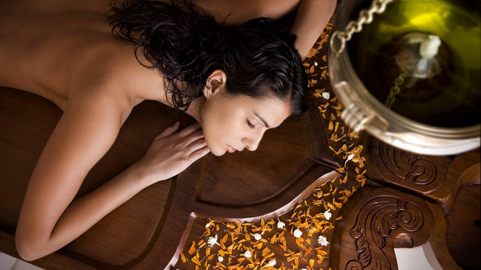 Side view of woman with bare back, shoulders lying on wood Spa table, orange flower petals