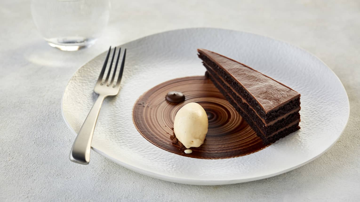 A thin sliced of a layered chocolate ganashe cake garnished with a dallop of cream on a white porcelain plate