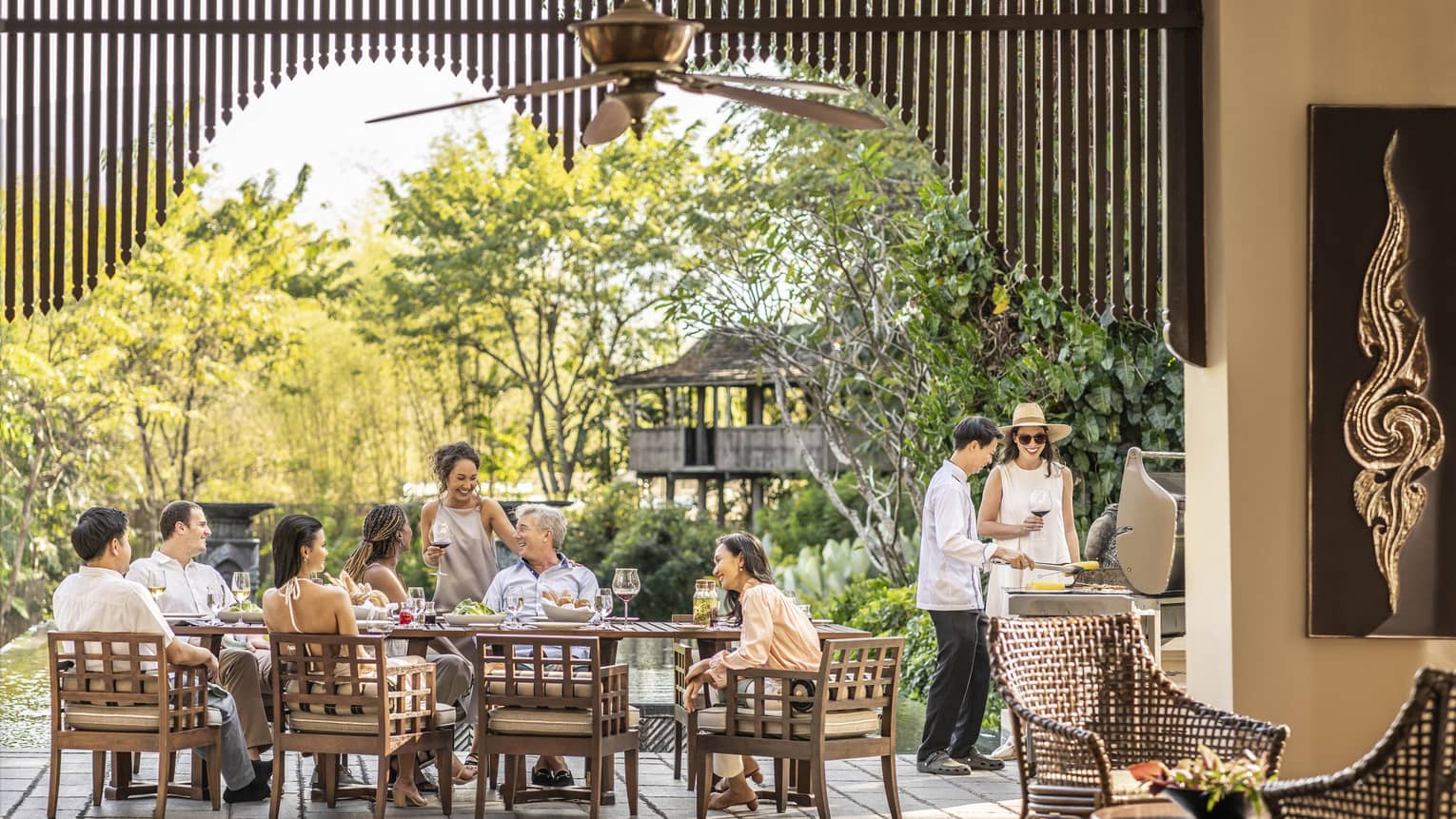 A long brown table with a family sat at it, in front of large trees.