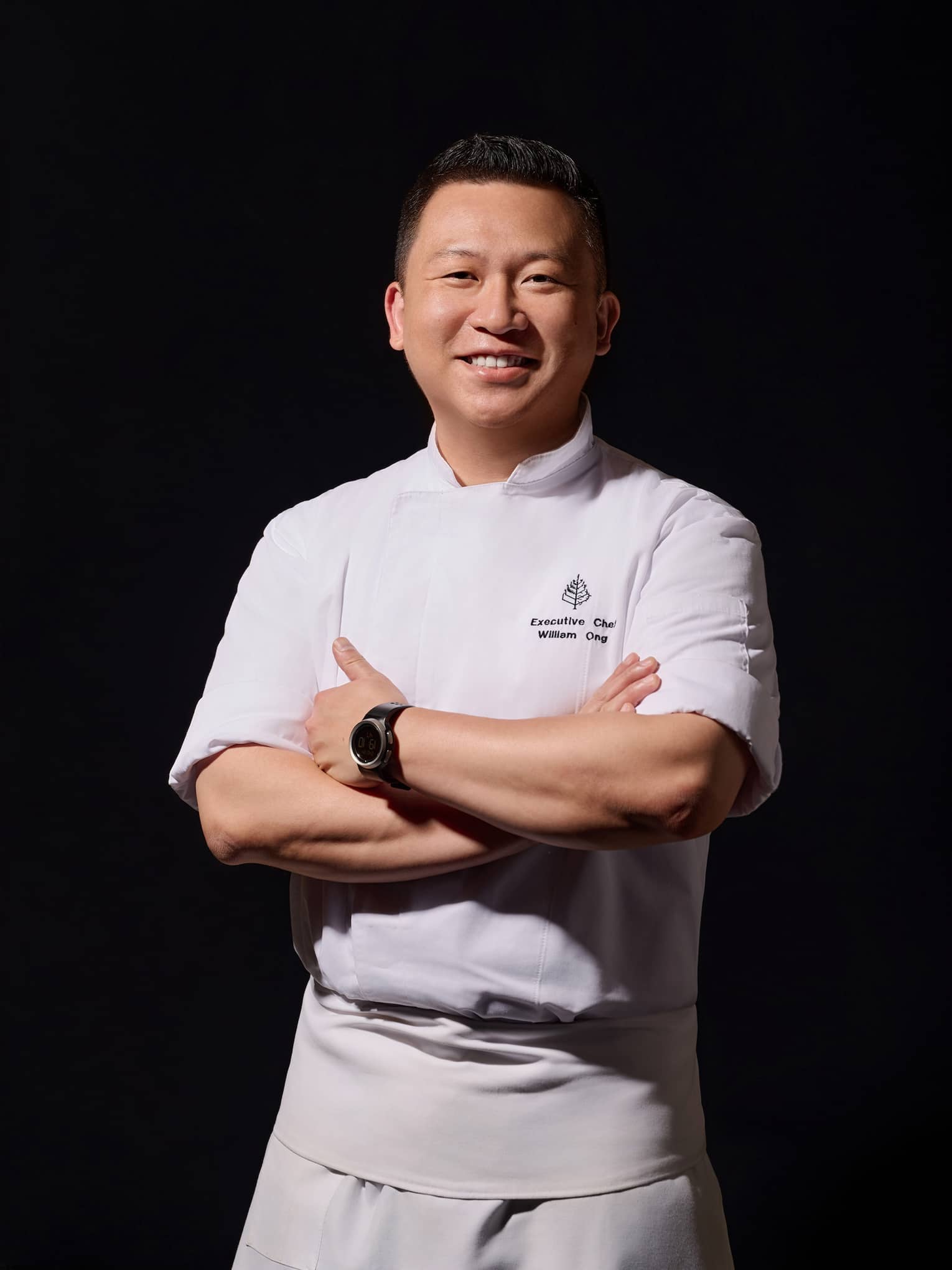 Smiling chef in chef?s coat with Four Seasons logo stands with arms crossed in front of black background