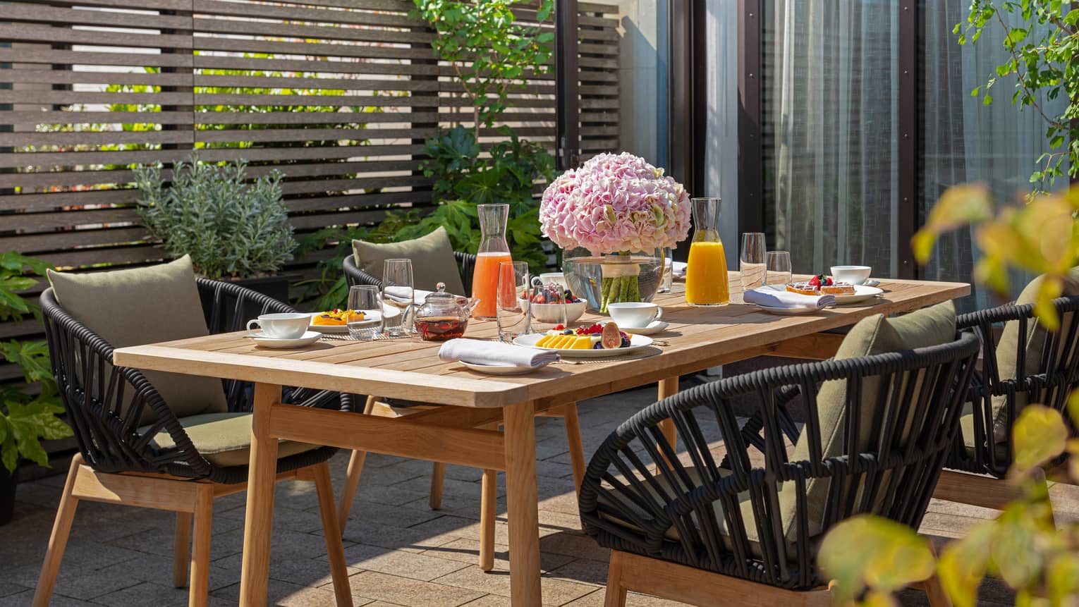 The Executive Conservatory at Four Seasons London Park Lane in Mayfair features an inviting outdoor dining area. A rustic wooden table is set with a colorful breakfast spread, including fresh fruits, pastries, juices, and tea. The table is beautifully decorated with a large bouquet of pink hydrangeas, enhancing the fresh, vibrant setting. Stylish black wicker chairs with comfortable cushions surround the table, nestled amidst lush greenery and a modern wooden slatted privacy screen. The scene is sunlit, offering a pleasant and serene dining experience.