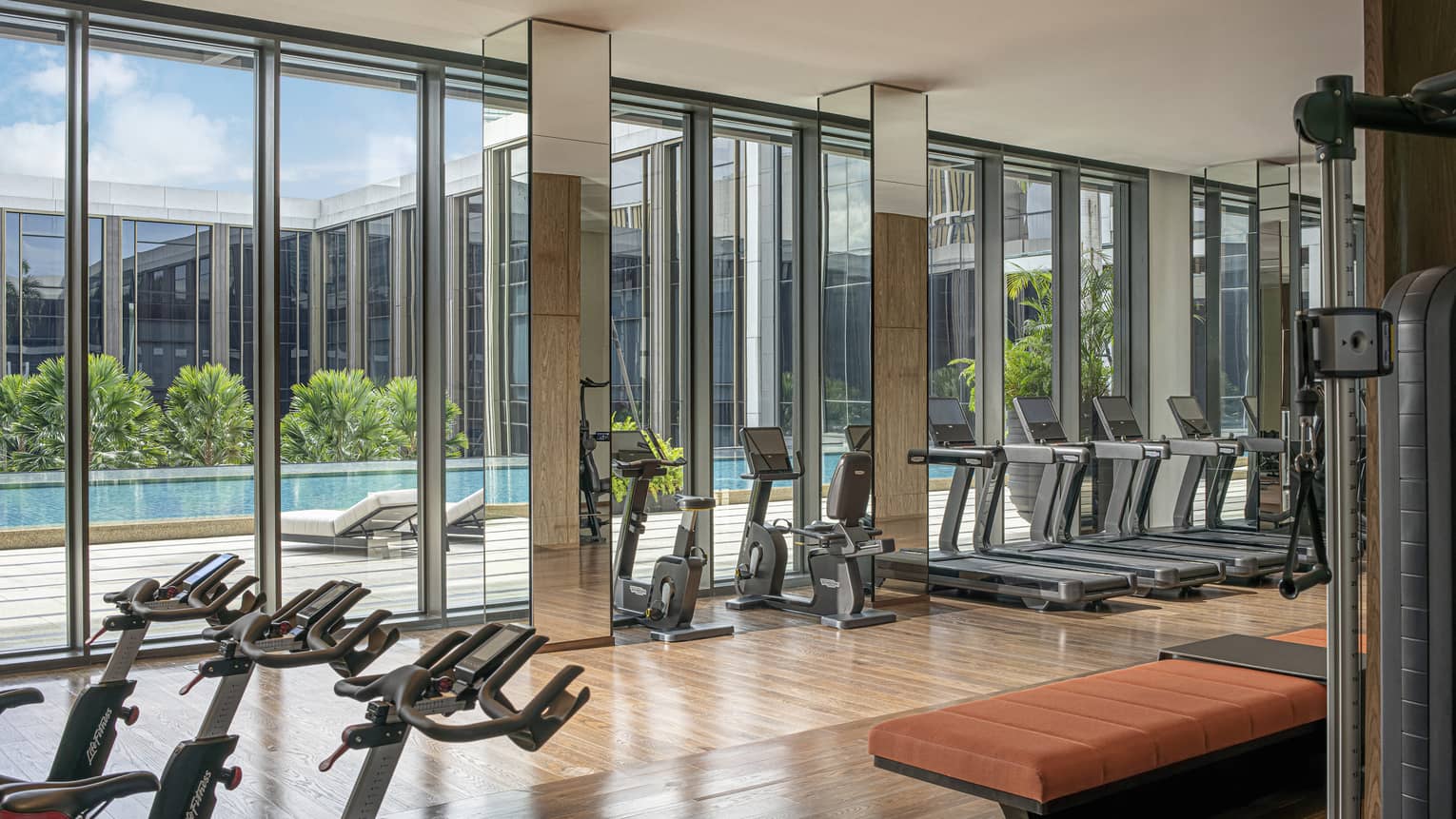Hotel gym with floor-to-ceiling windows and rows of exercise equipment, natural light, pool views