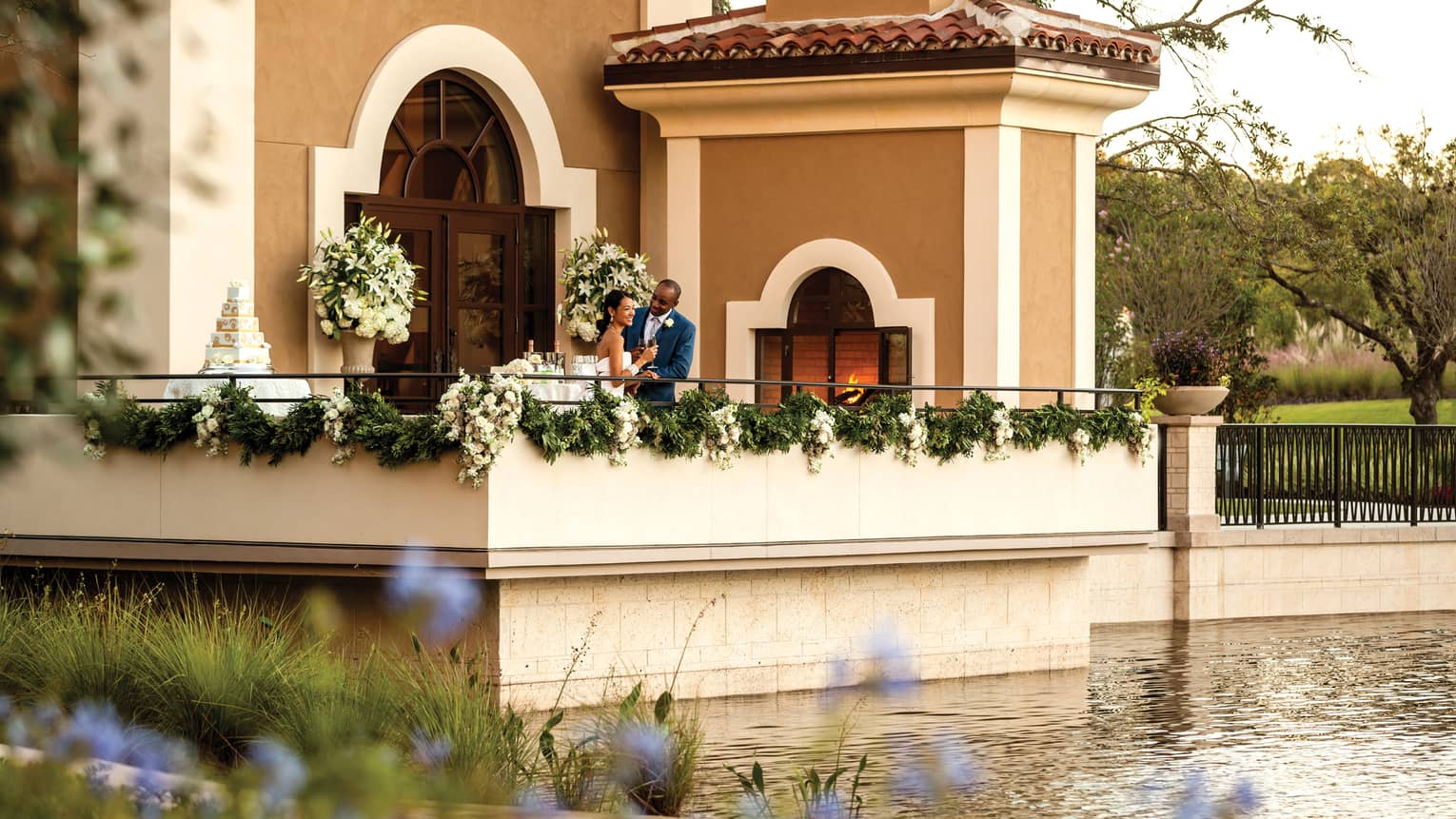 View across water to patio with bride and groom with Champagne, wedding cake, door to event space