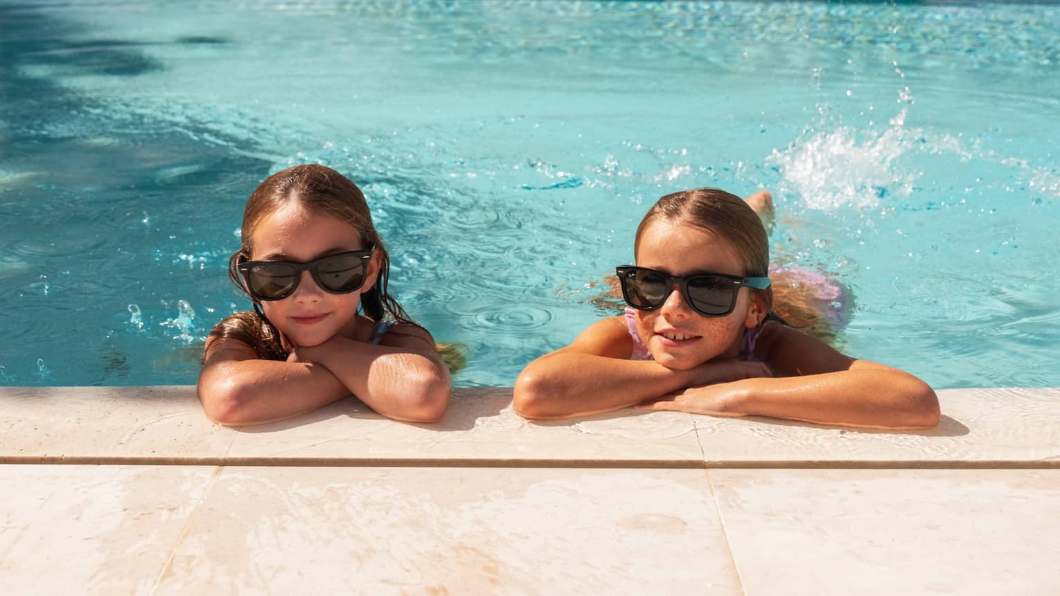 Two kids wearing sunglasses and swimming in the pool.