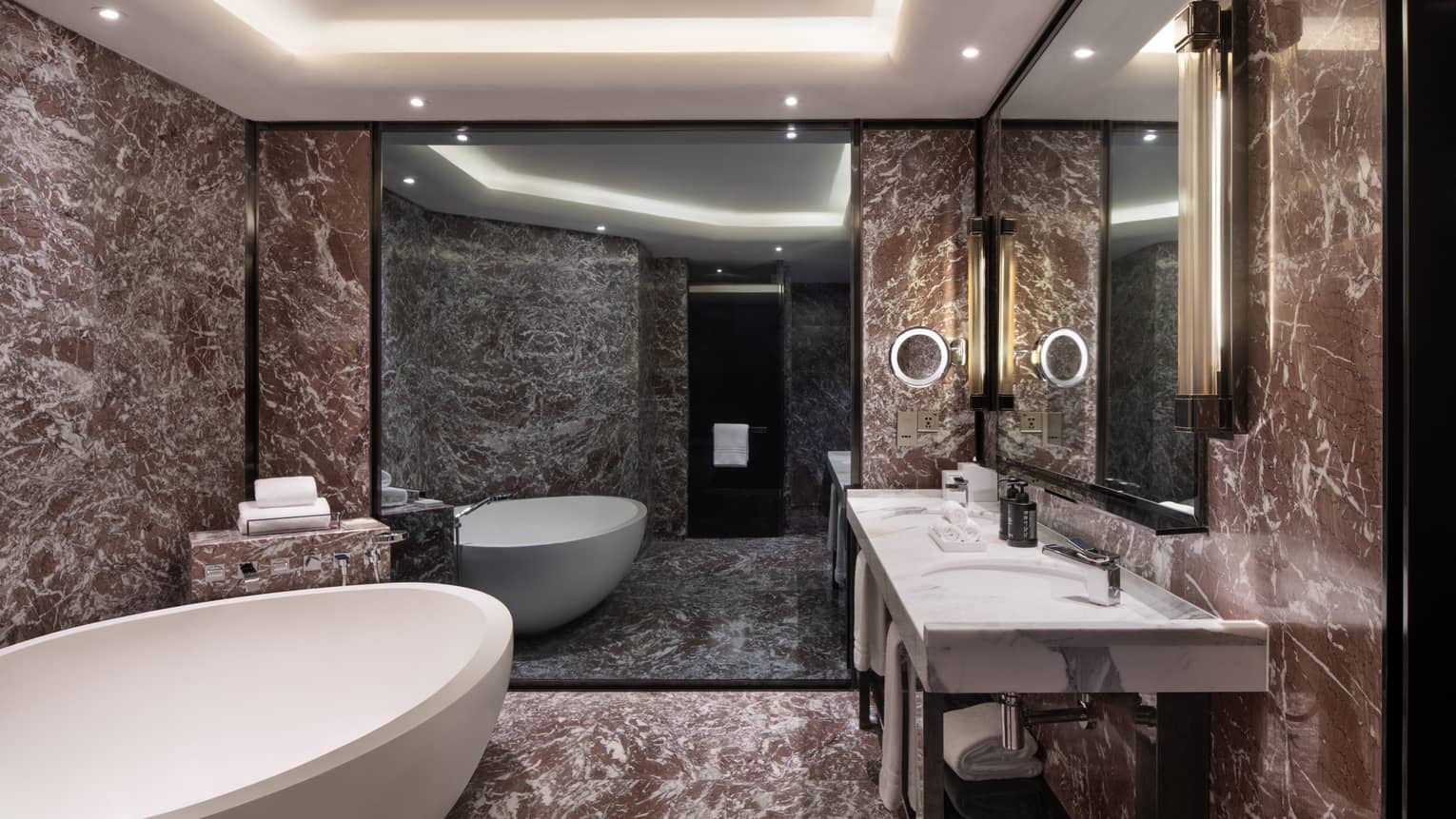 Guest room bathroom with chocolate marble accents, large soaking tub, double vanity with lighted wall sconces