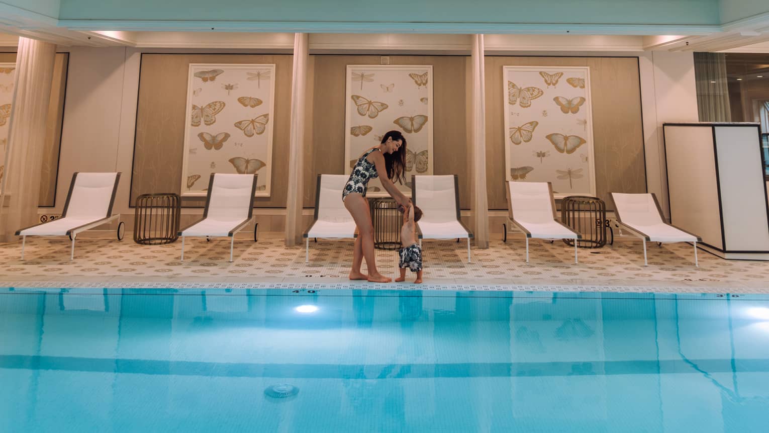 A woman and child standing by an indoor pool and lounge chairs.