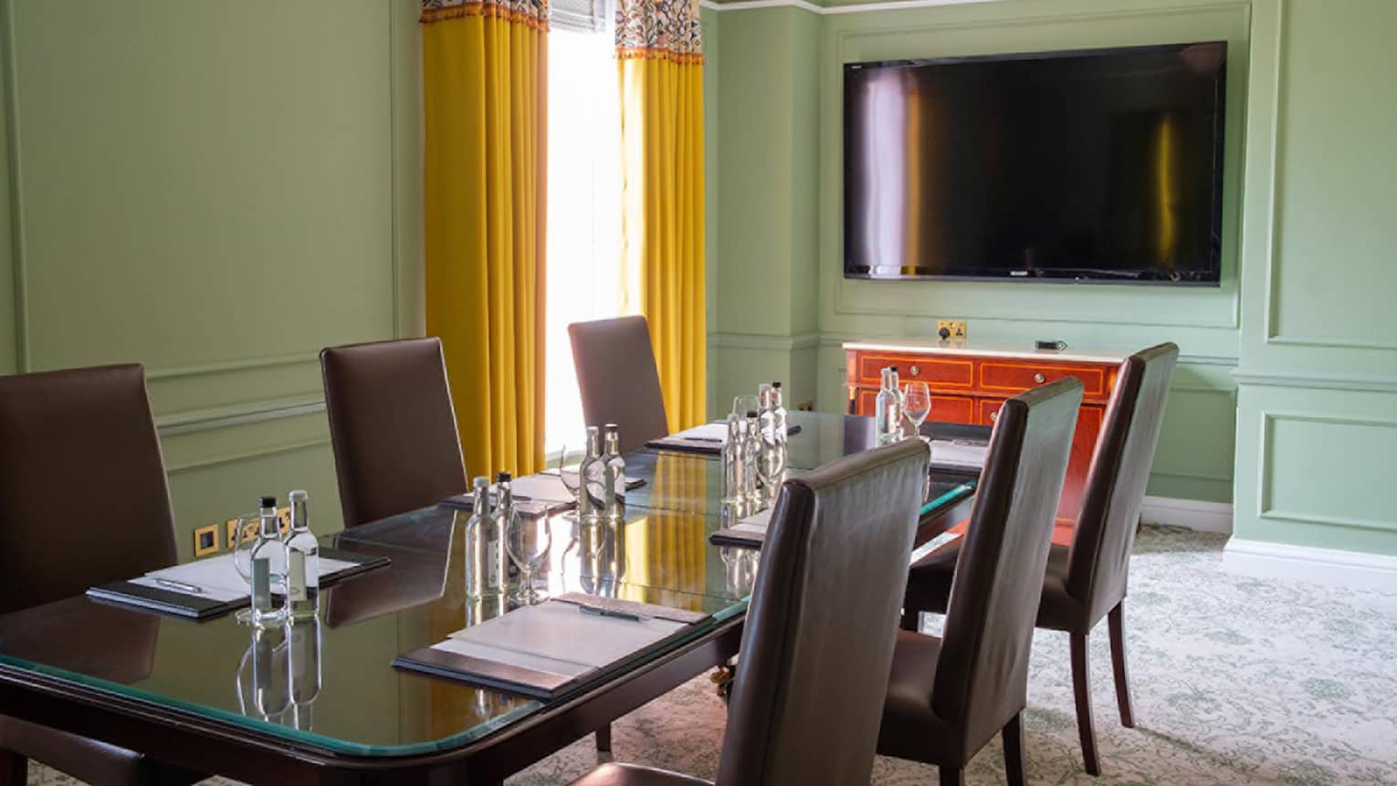 Green-walled boardroom with glass-topped meeting table, brown leather chairs and wall TV