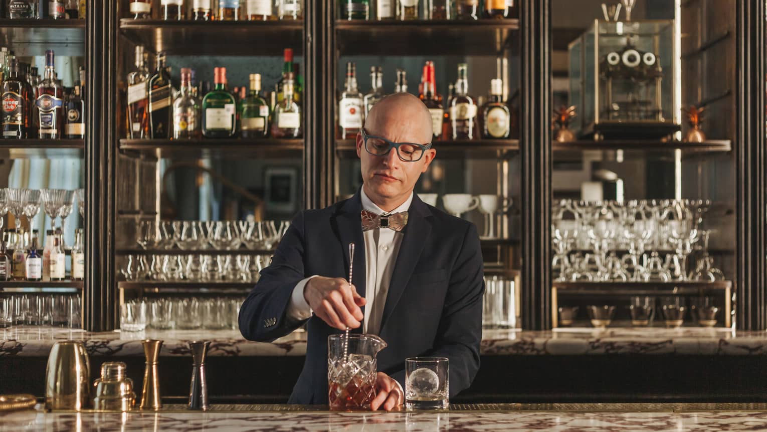 A bartender in a suit and bowtie stirring a drink with cabinets of liquor behind him.