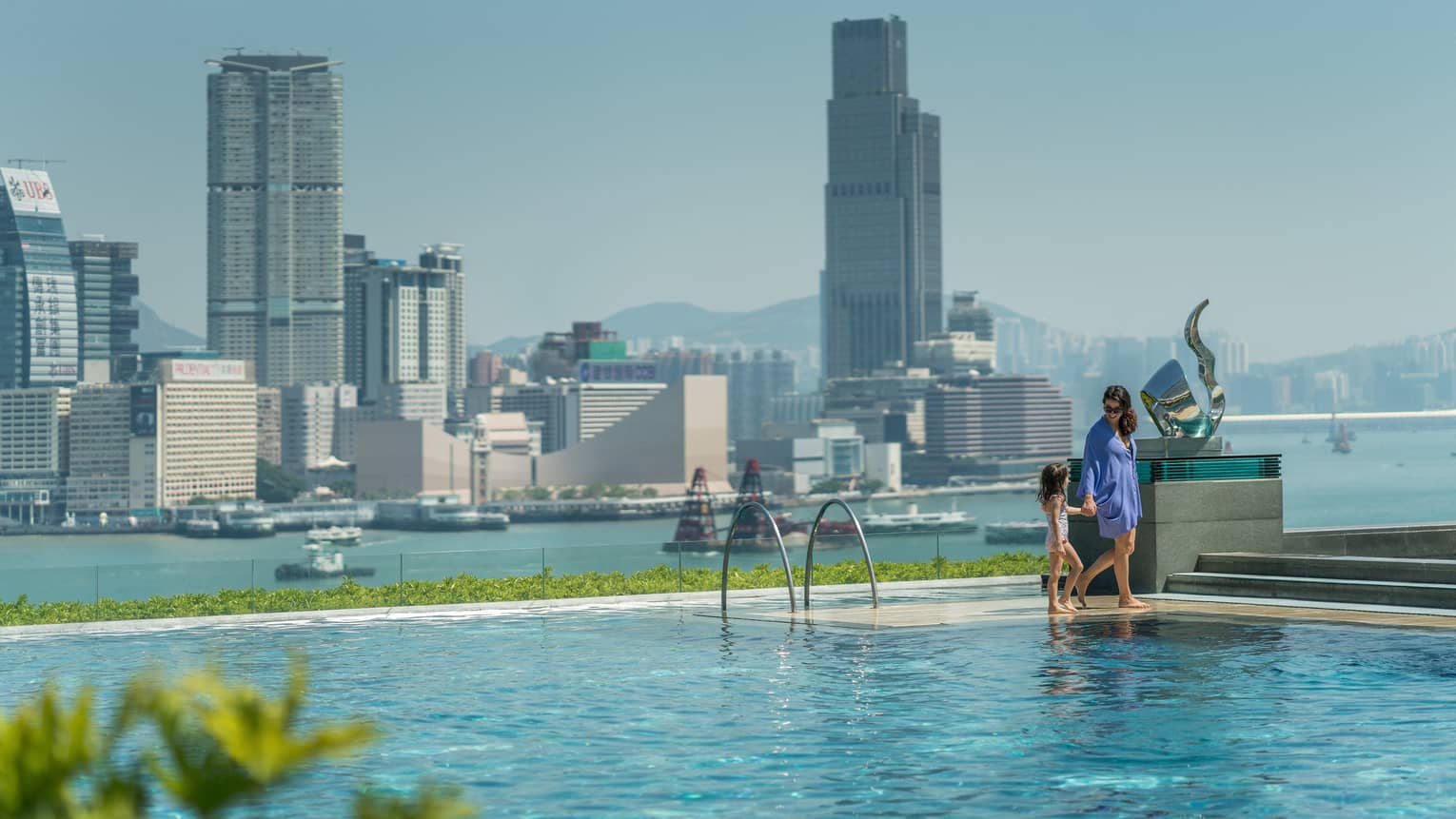A parent and child walking along the edge of an infinity pool overlooking a skyline