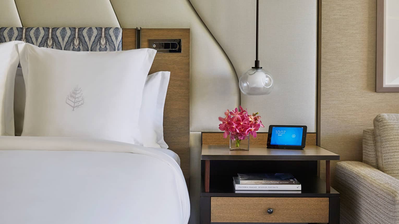 Hotel room with white bed, Four Seasons logo on pillow, large white padded headboard, flowers and ipad on nightstand