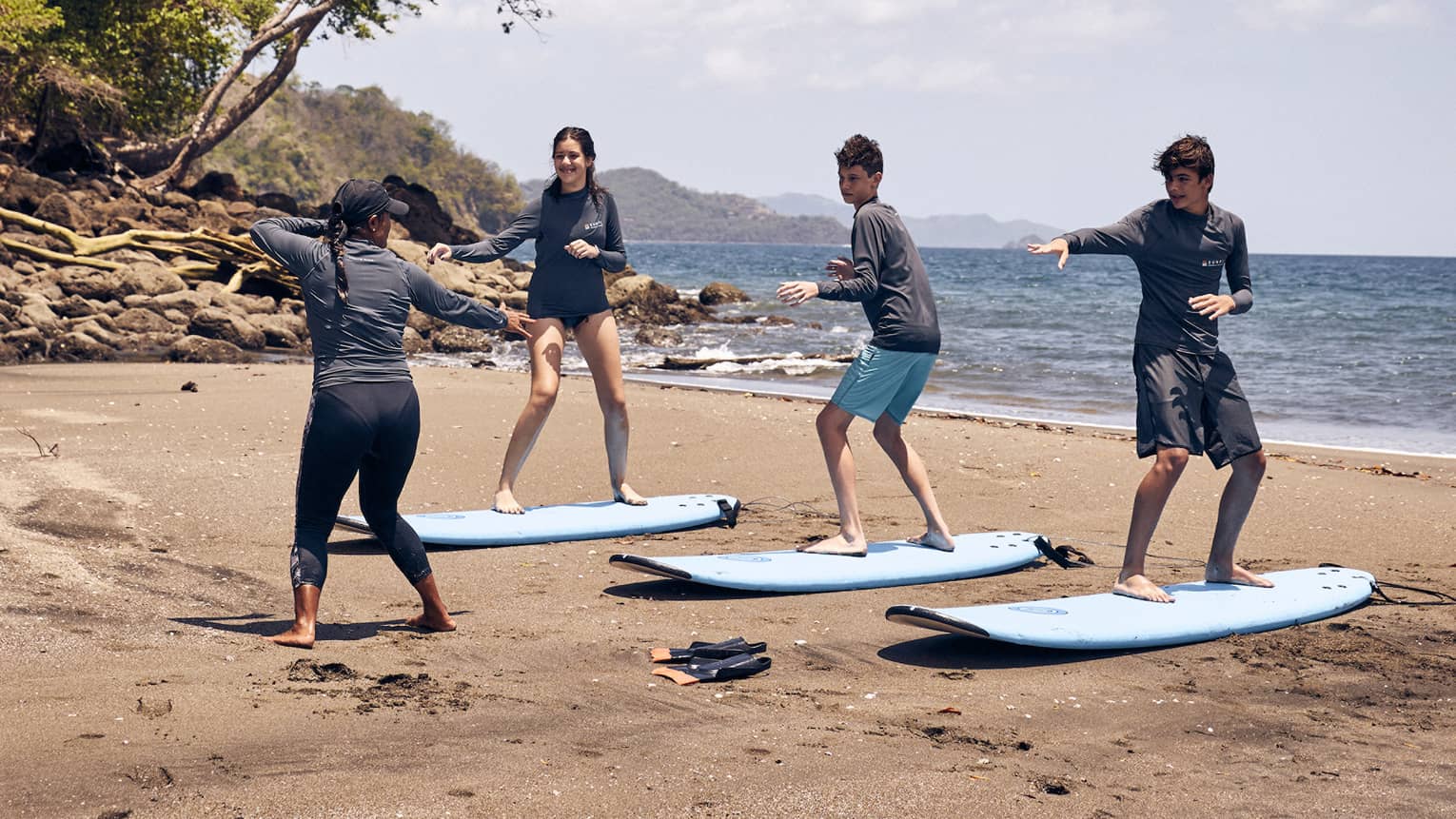Three young people practice standing on a surfboard on the sand while an instructor guides them