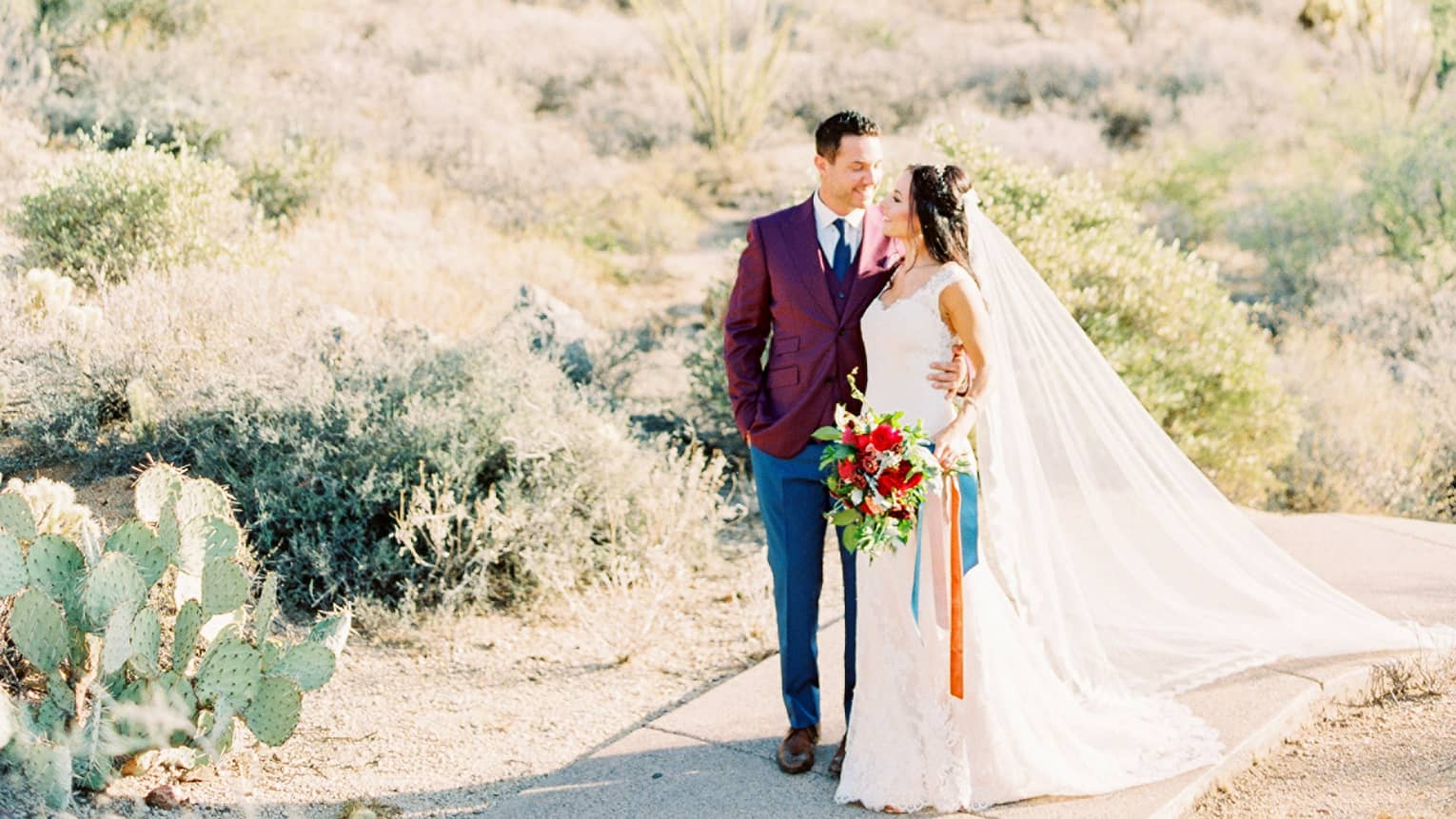 Groom and bride with colourful floral wedding bouquet pose in desert near cactus
