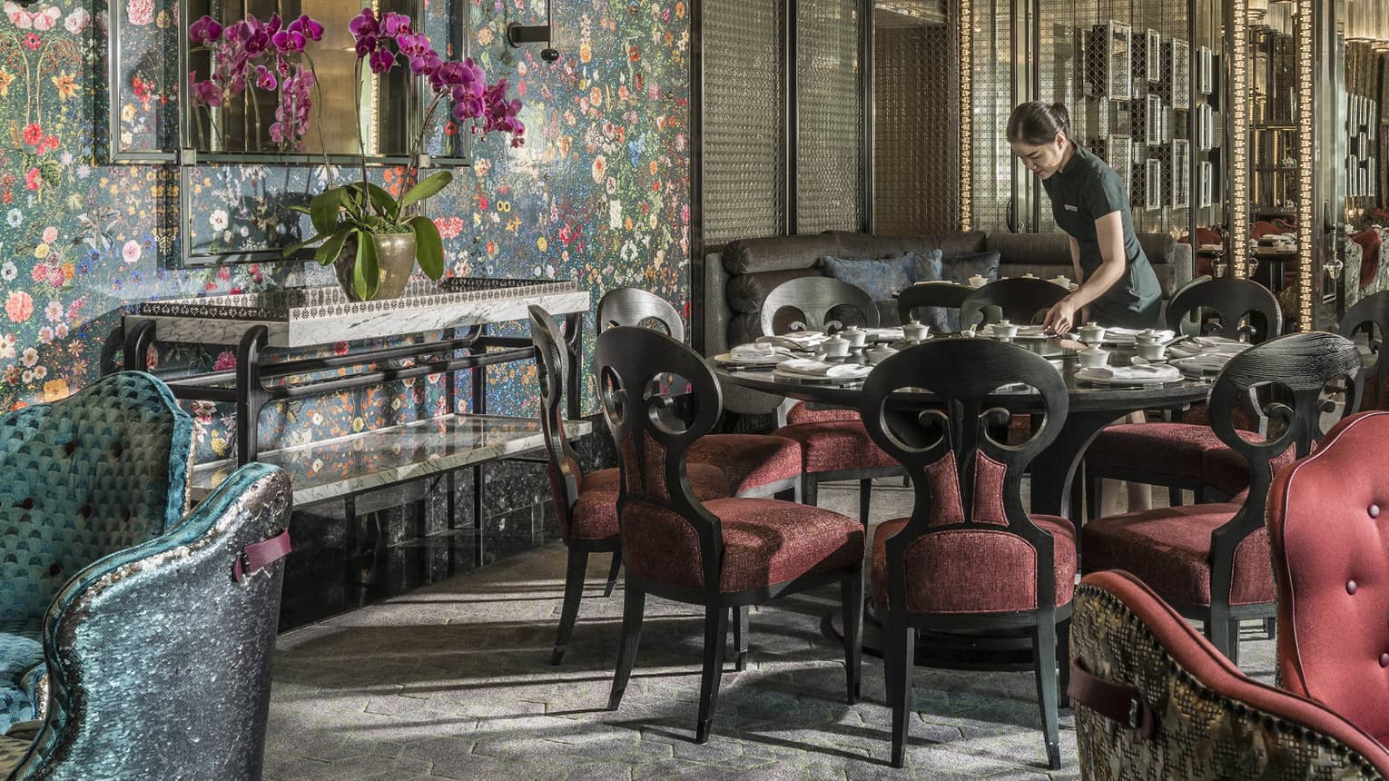 Hotel staff sets table with elegant red velvet chairs under tall mirror, floral wallpaper 