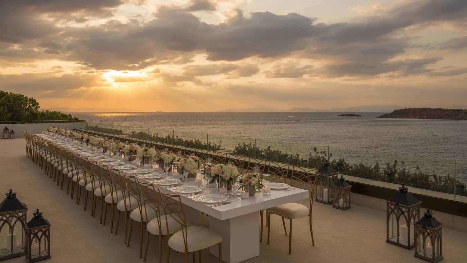 Long white table with flowers, white and gold chairs, surrounded by lanterns by ocean