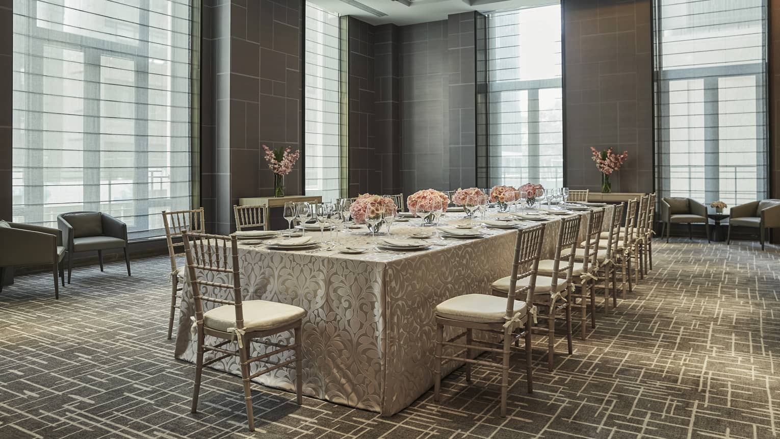 ,A long table with table cloth and flower centerpieces in a room with large windows.