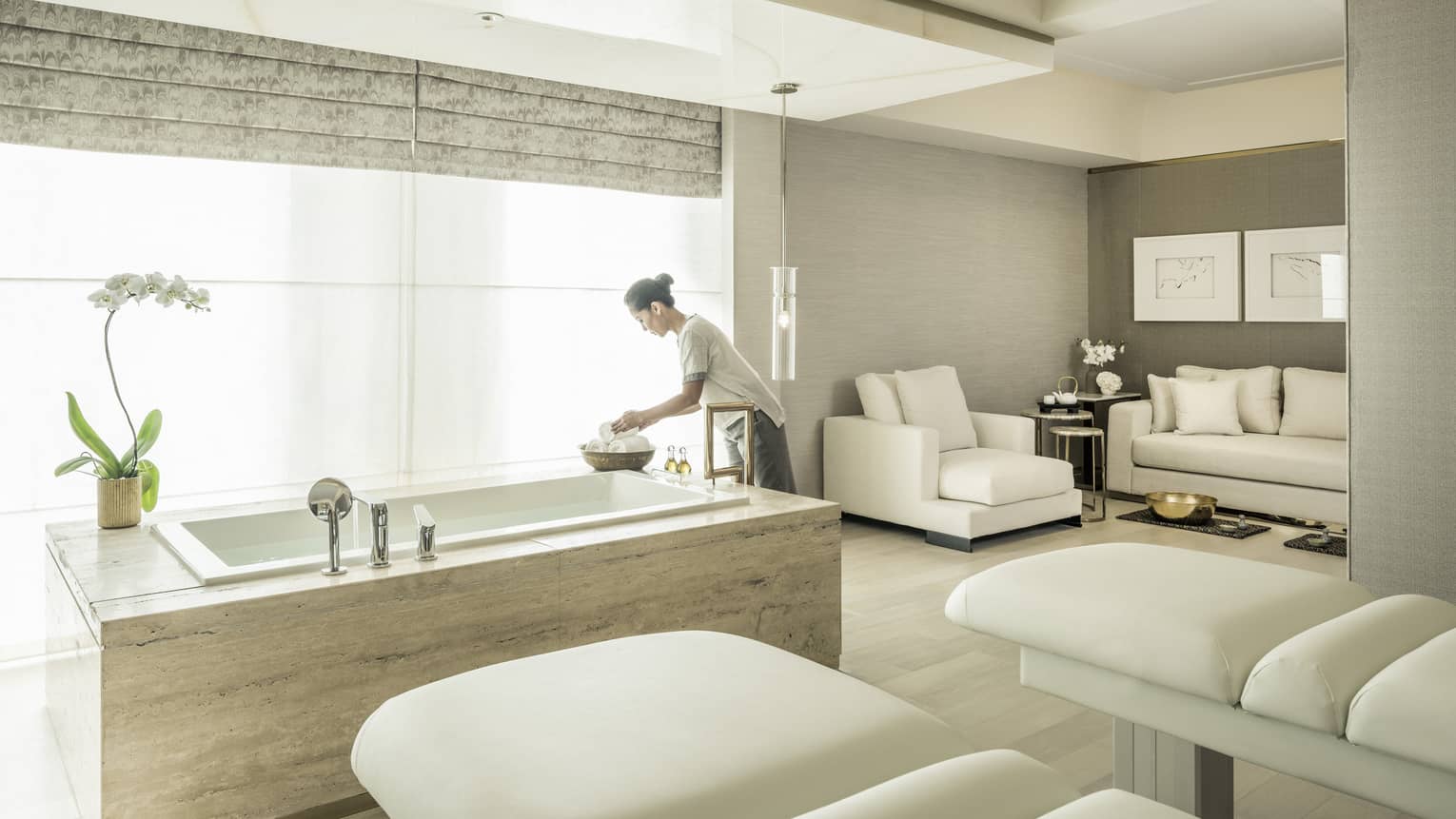 A spa attendant arranges some products in the spa suite, with two massage beds and a soaking tub in front