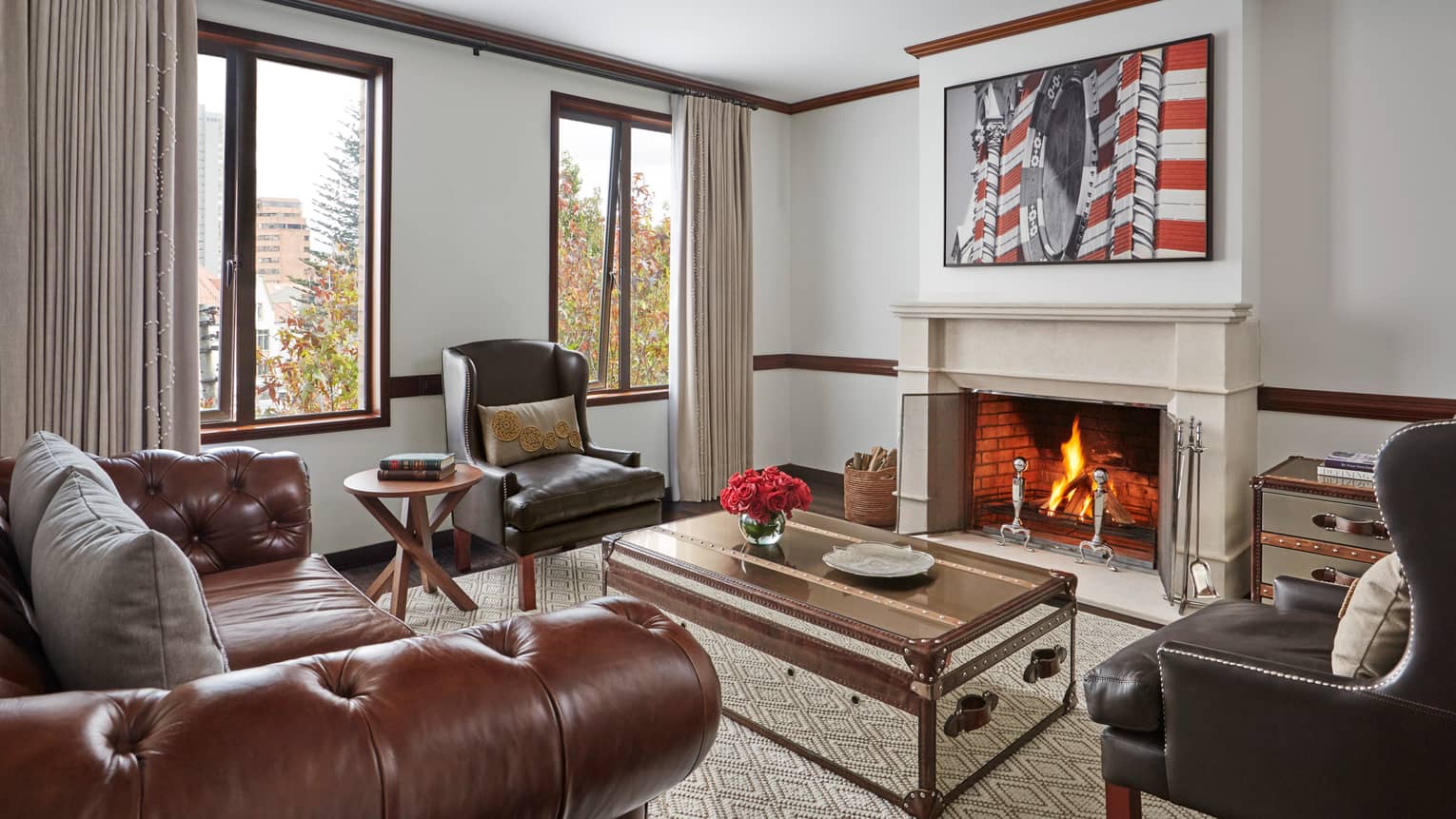 Hotel living room with brown leather sofa and armchairs trunk-style coffee table in front of roaring fireplace