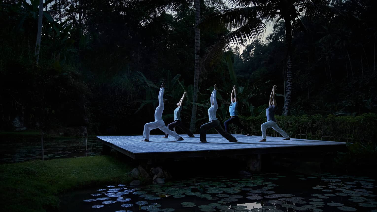 Five people kneel in yoga pose with arms above head on wood platform in forest at night