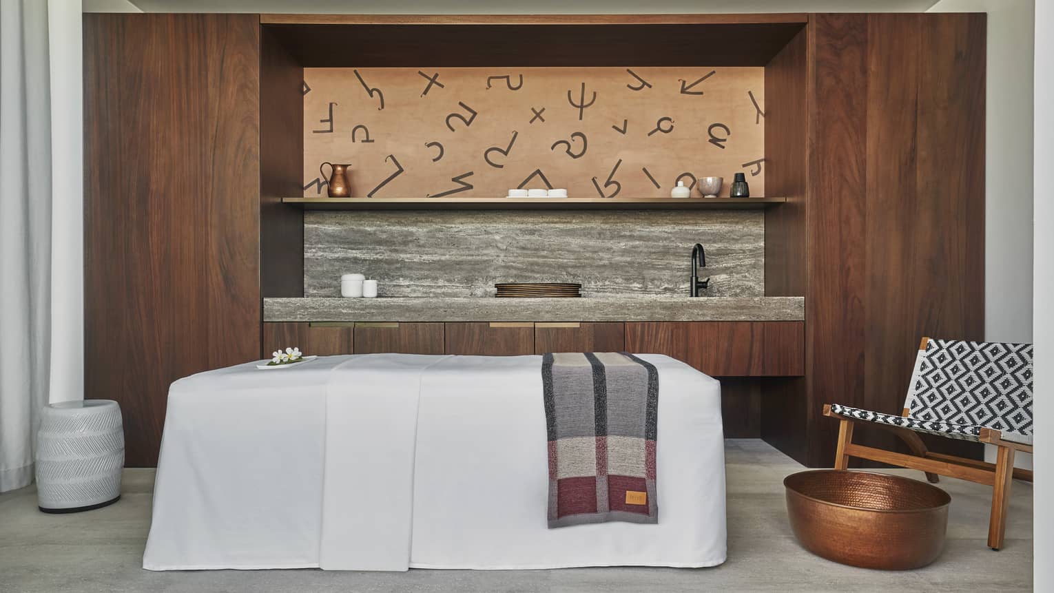 A Four Seasons Resort Los Cabos spa room is decorated with a walnut wooden accent wall with a gray concrete shelf, a mid century modern chair with a blue and white tribal pattern and a massage table covered with a white cloth 