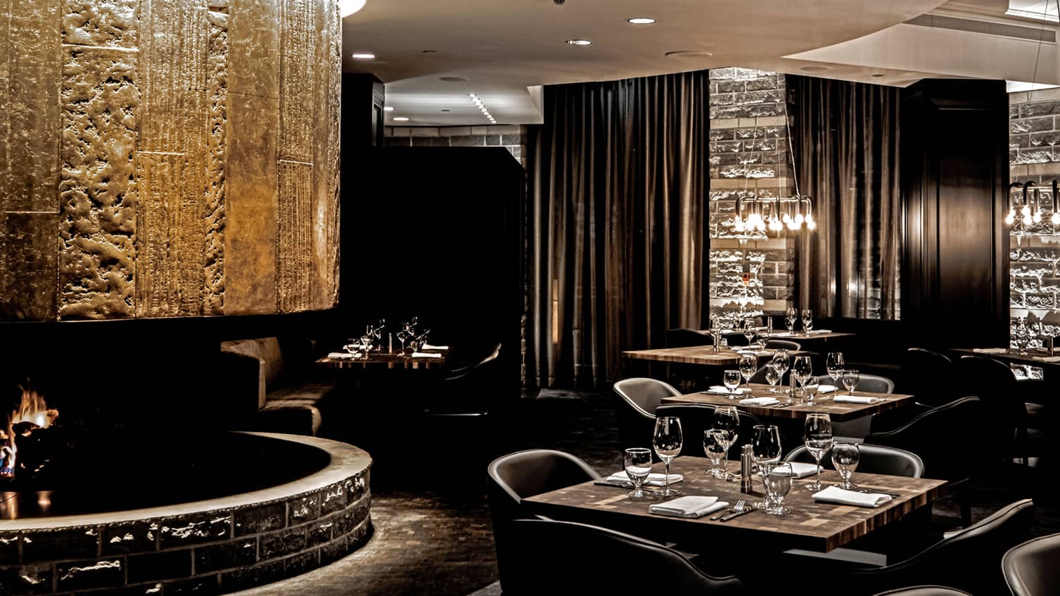 Newly renovated steakhouse with dark wood tables set for four, black curtains, lowlights, fireplace 