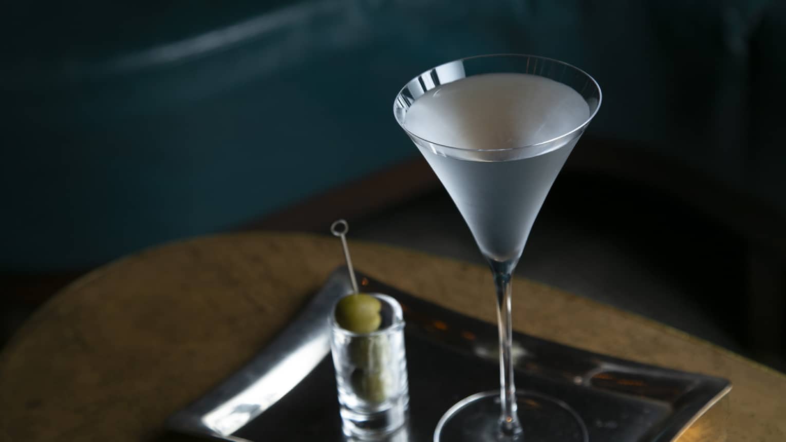 Signature cocktail in stemmed glass on silver tray, skewer of olives