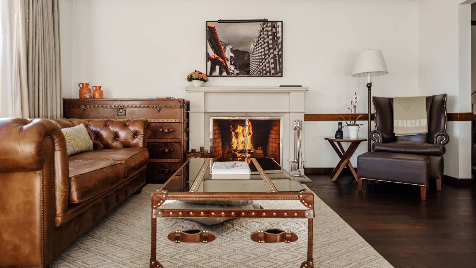 A brown leather chesterfield sofa, dark-brown leather arm chair and two trunk-style tables frame a central fireplace in the living room