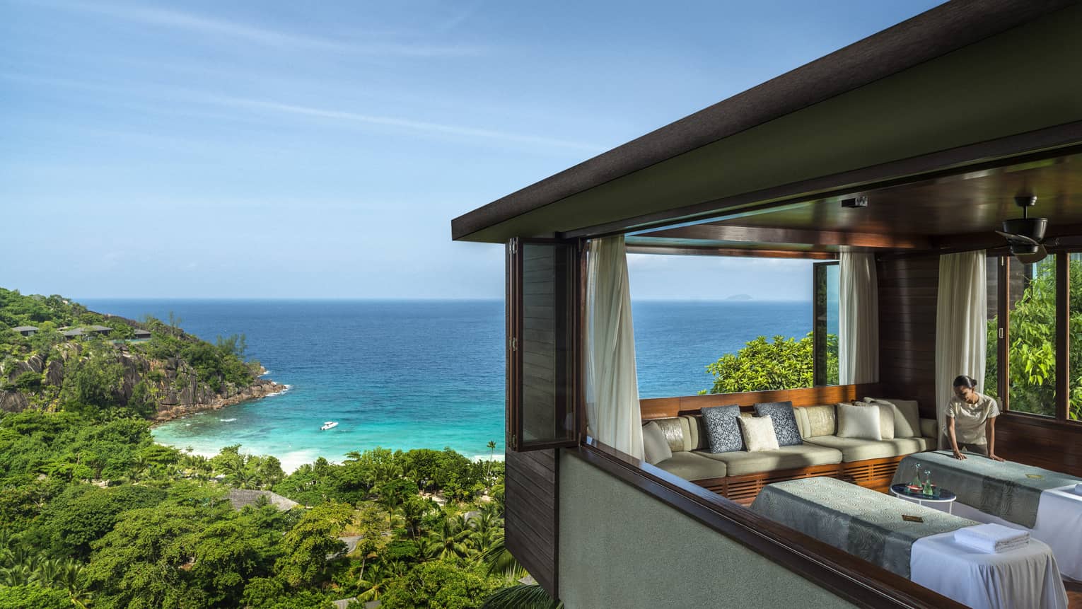 Woman smoothes sheet on side-by-side massage tables in spa room high above trees, ocean