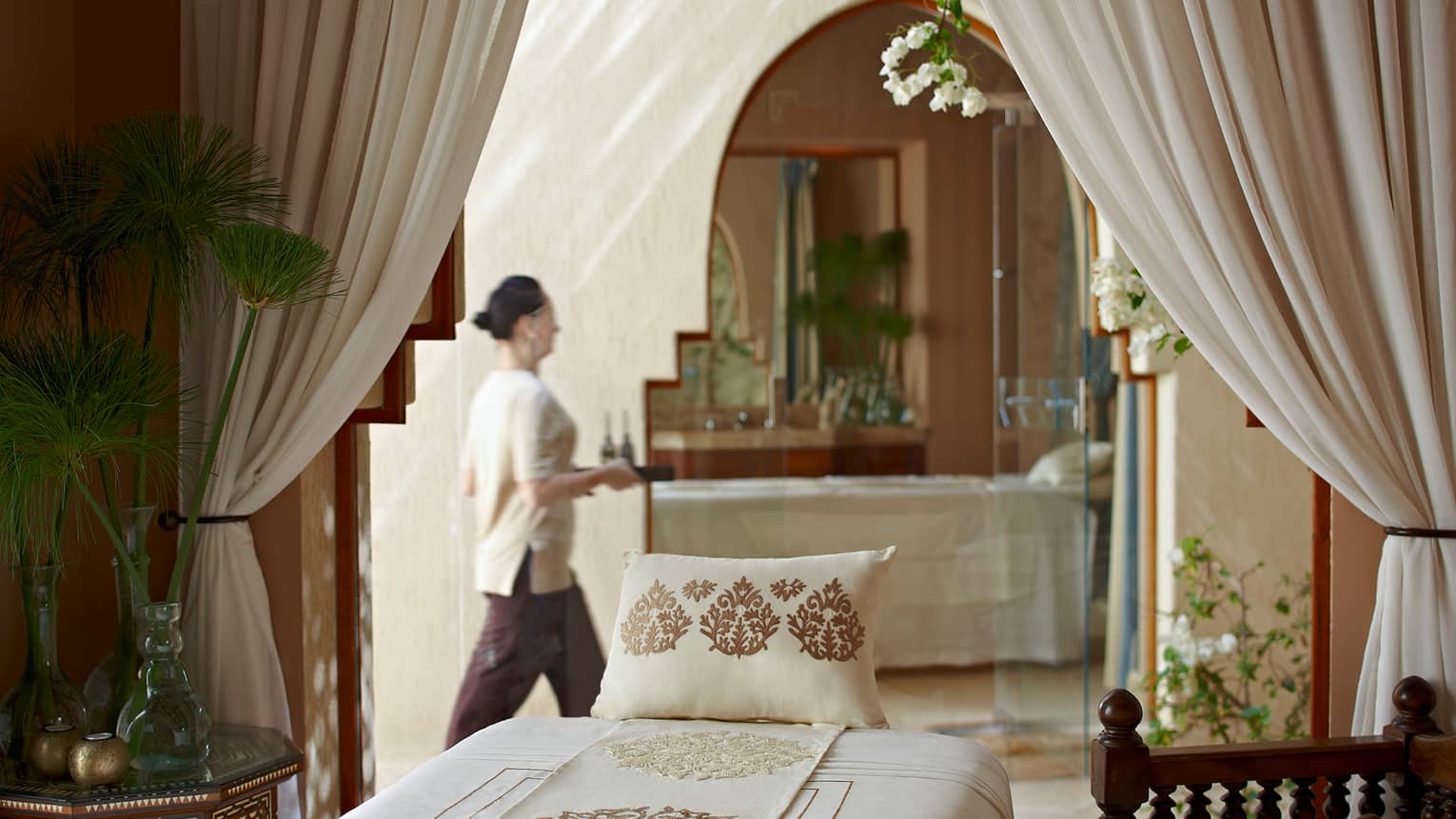 Woman with tray walks behind massage bed with embroidered pillow in spa