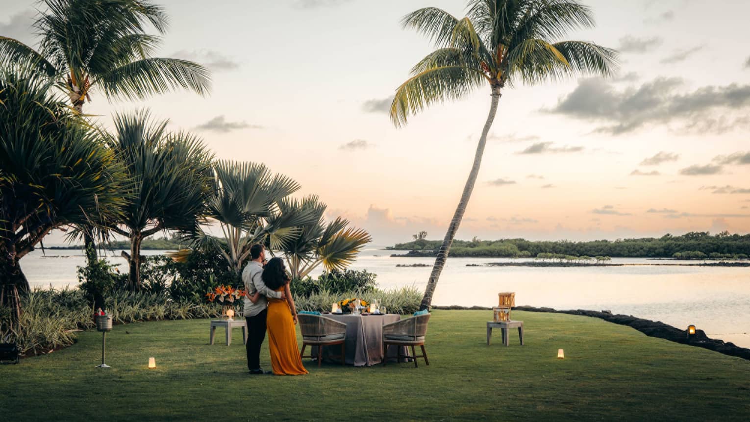 A couple stand on the shore, arms around each other, gazing toward the ocean; set table with two chairs beside swaying palms.