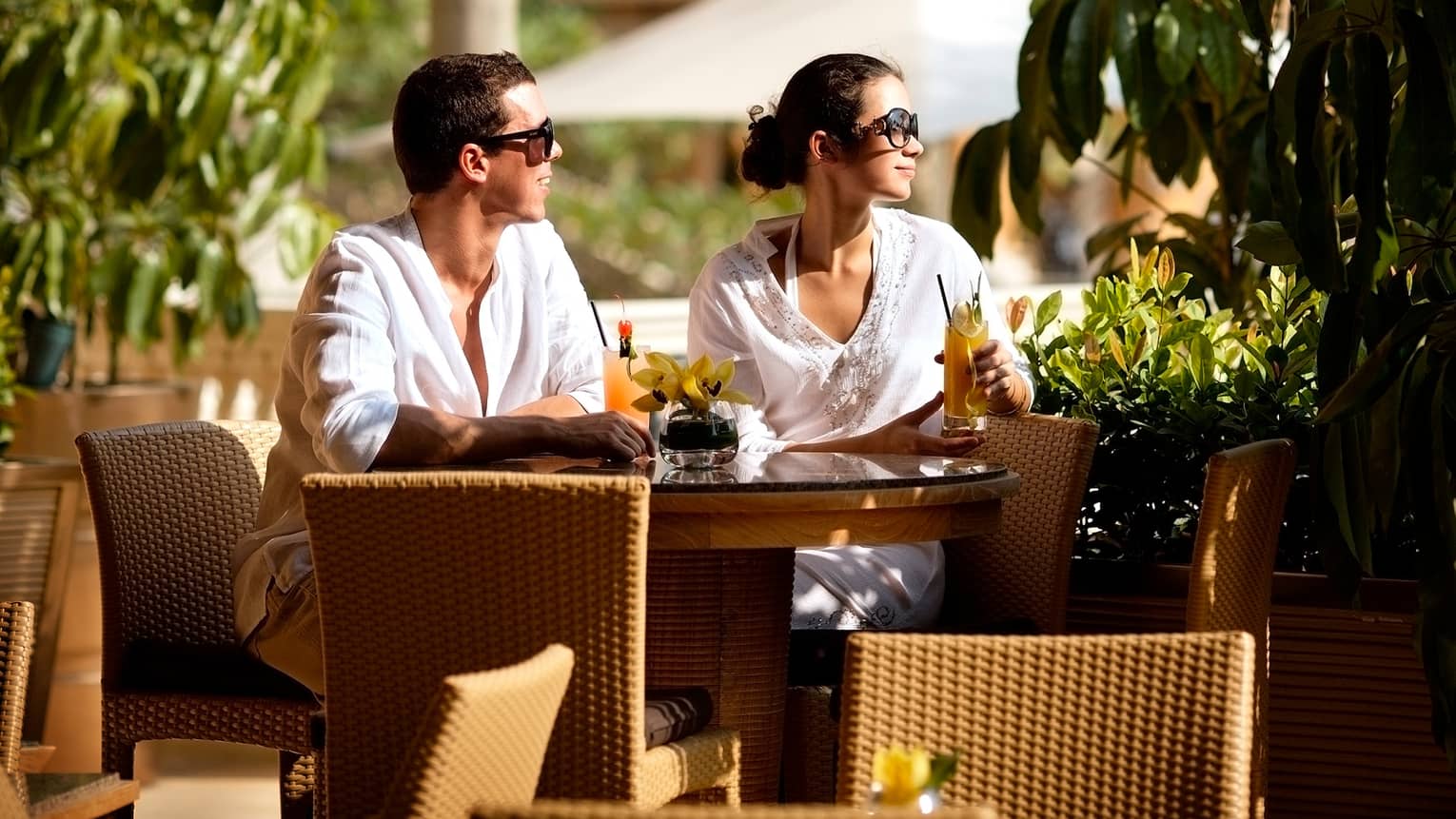 Couple wearing white linen shirts, sunglasses drinks cocktails at patio table