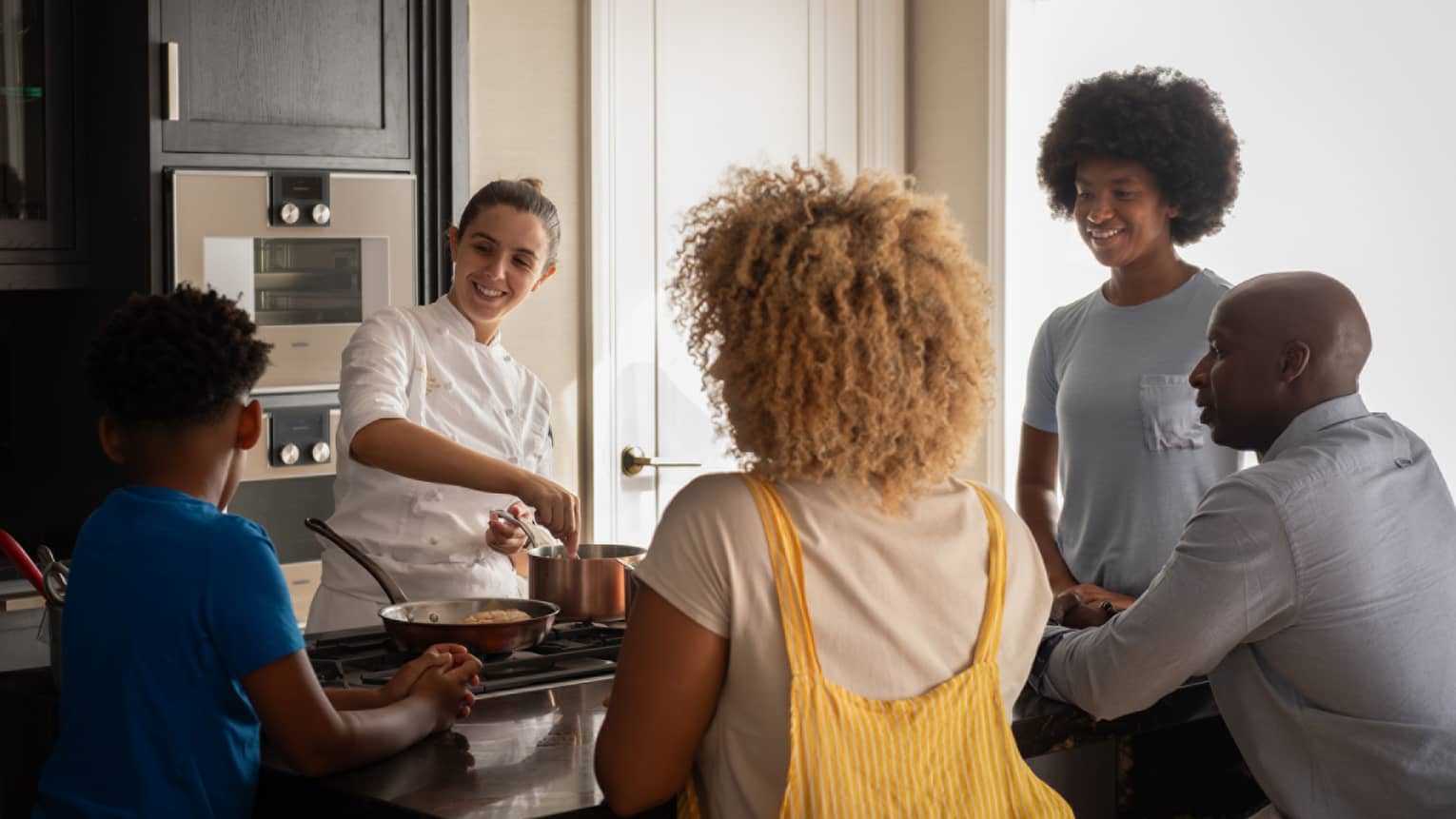 A family of four gathers around a kitchen-island cooktop watching a smiling chef prepare food in copper cookware. 
