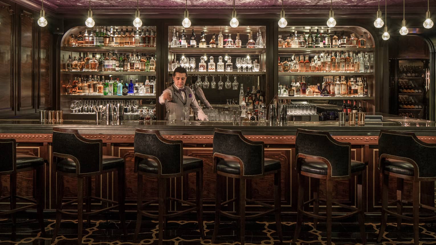 SIRR bartender in vest stands behind bar stirring cocktail, glasses and bottles lining wall behind him