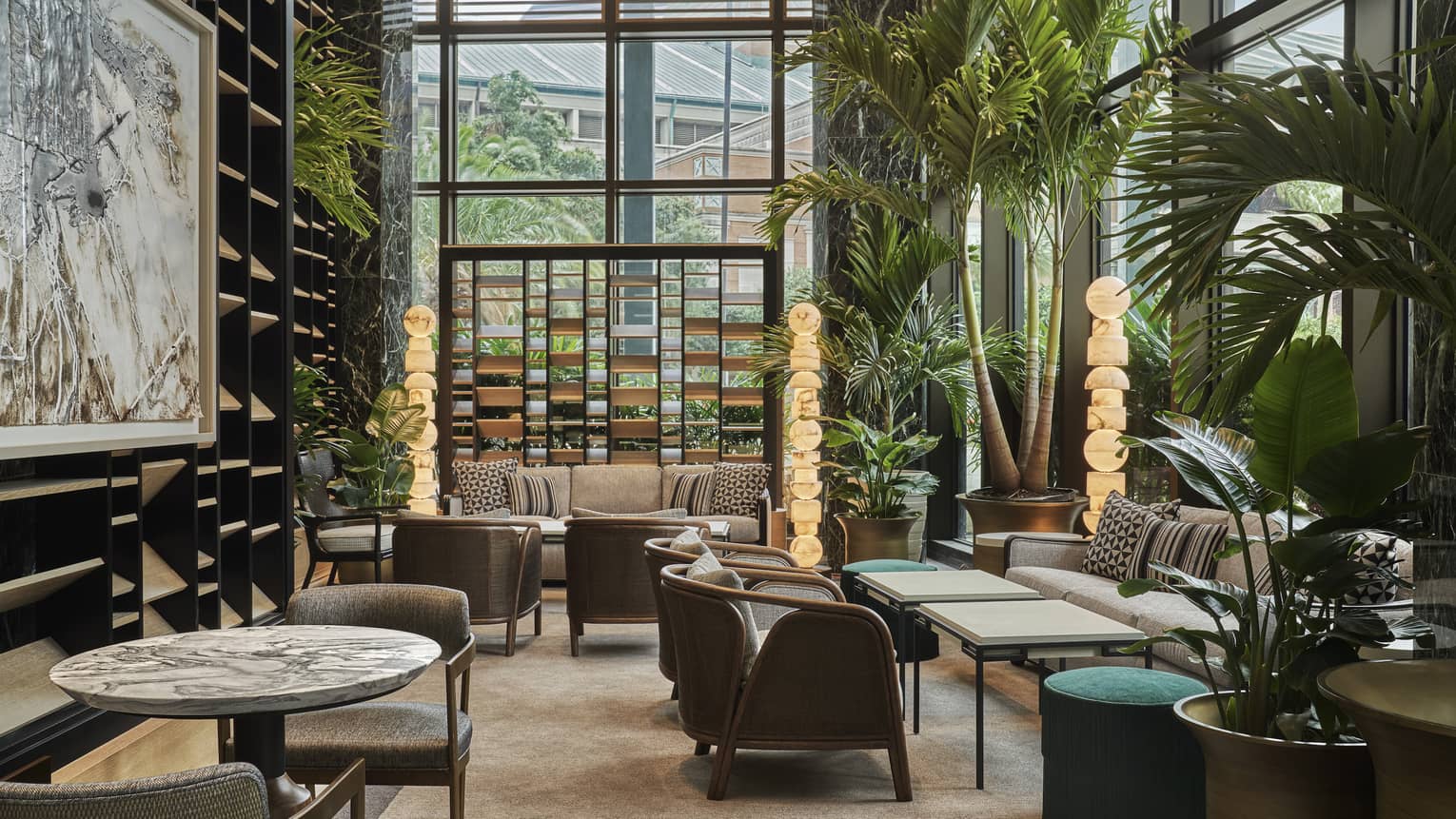 Chandelier Bar seating area with brown accent chairs, marble-topped tables, floor-to-ceiling windows and potted palms