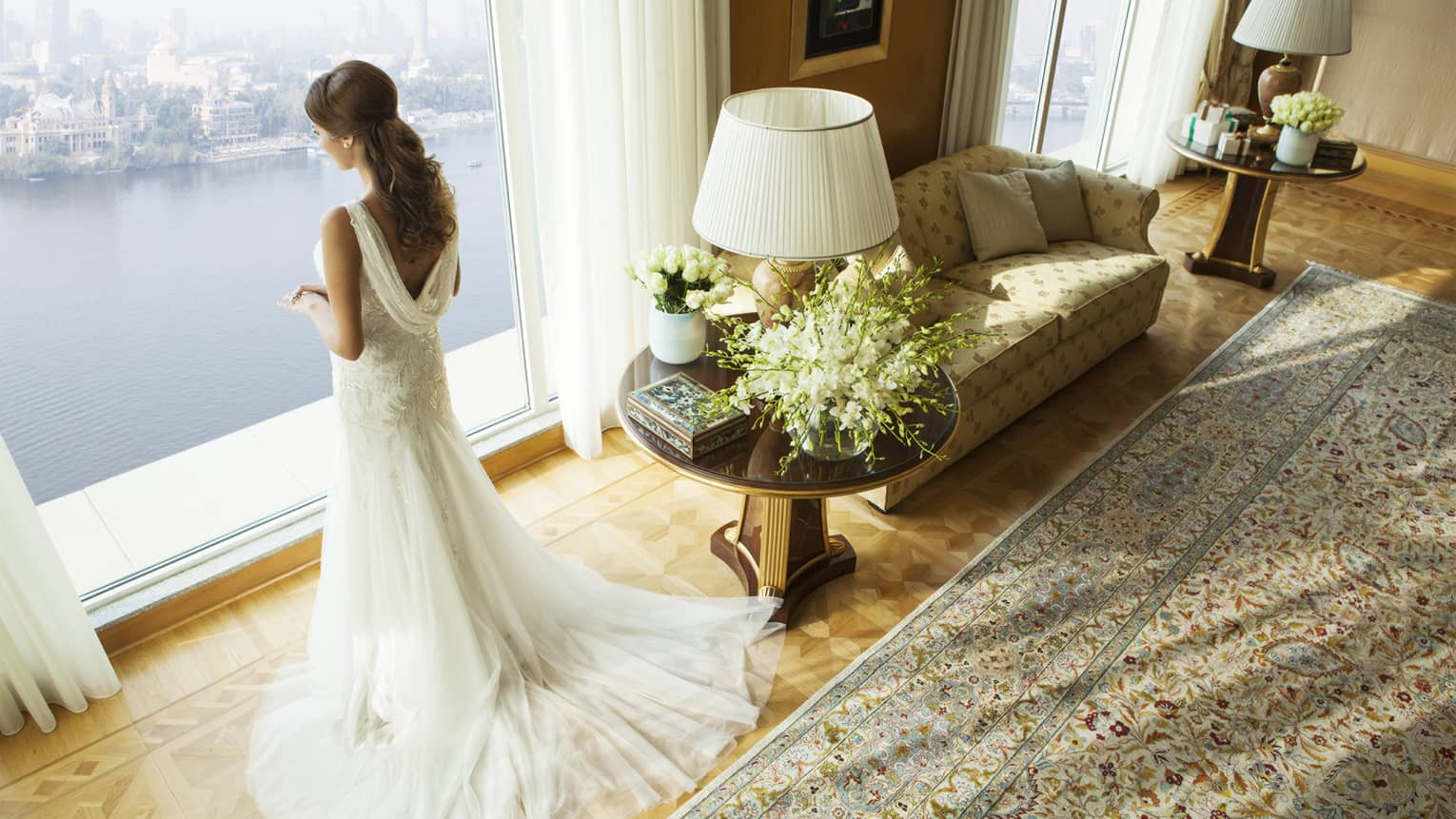 Back of bride with long white wedding gown and train, standing beside sofa in Royal Suite and looking out window