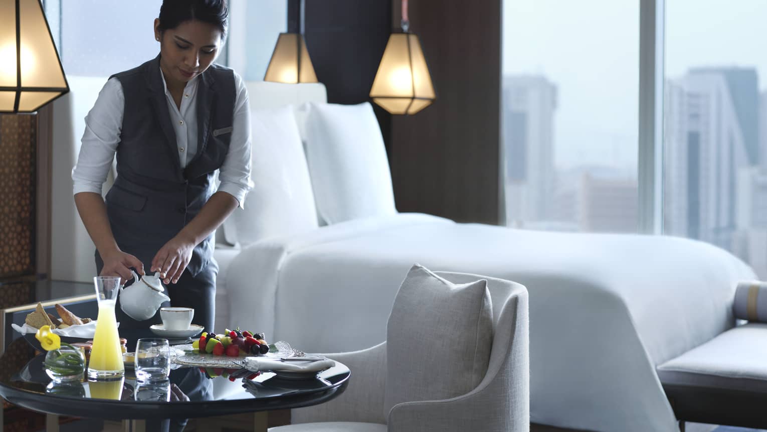 Woman in hotel suite pours tea into a white teacup next to a plate of fruit and bread.
