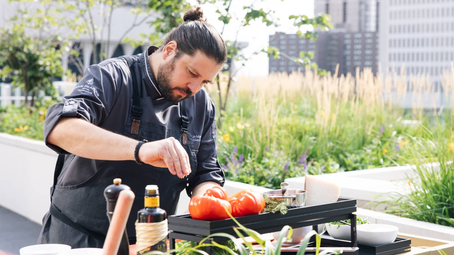 Chef Martin Morelli wears a black chef's jacket and apron as he seasons sliced tomatoes on an outdoor table surrounded by other ingredients