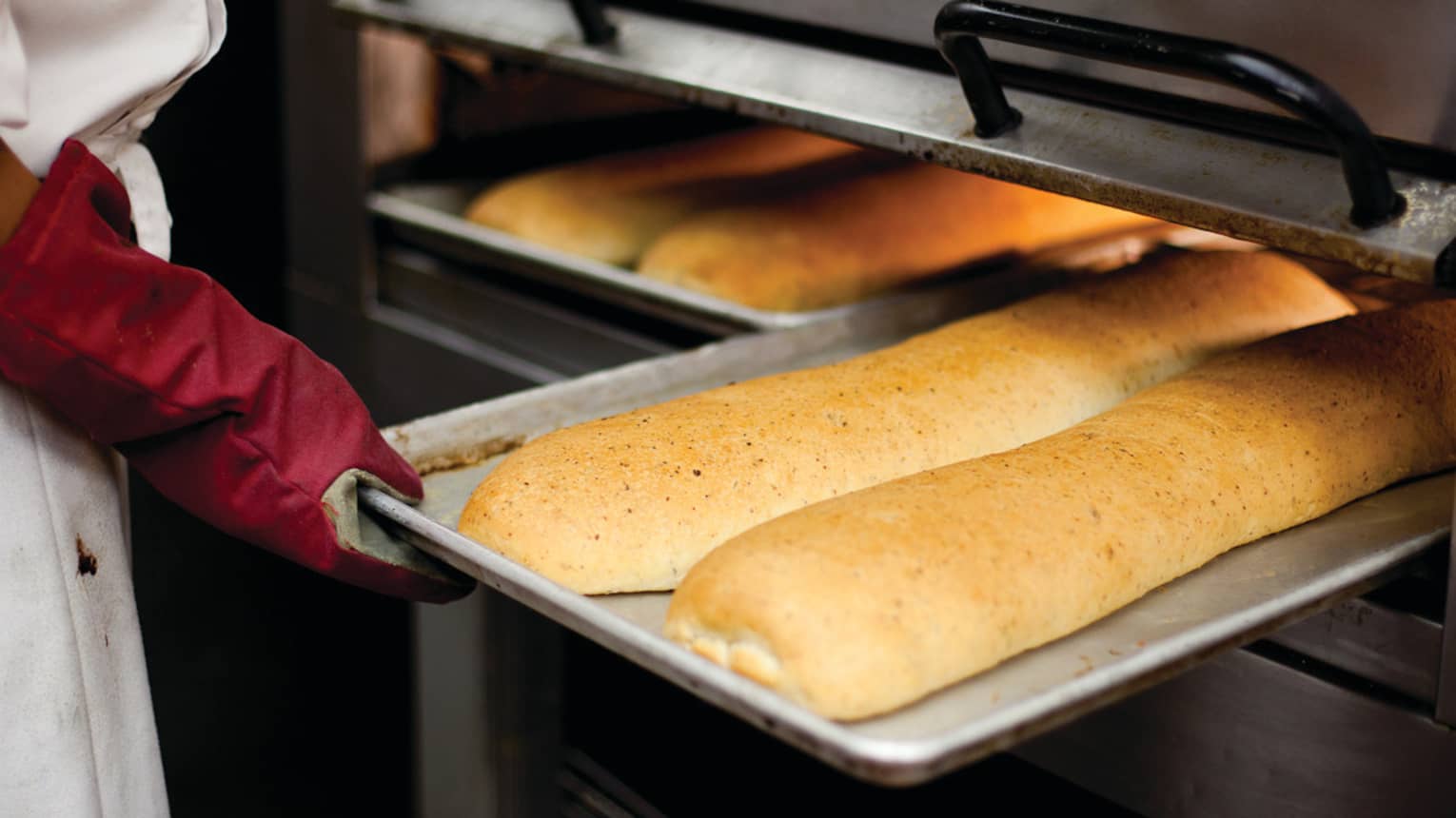 Chef wearing oven mitt removes tray with fresh baked bread from oven