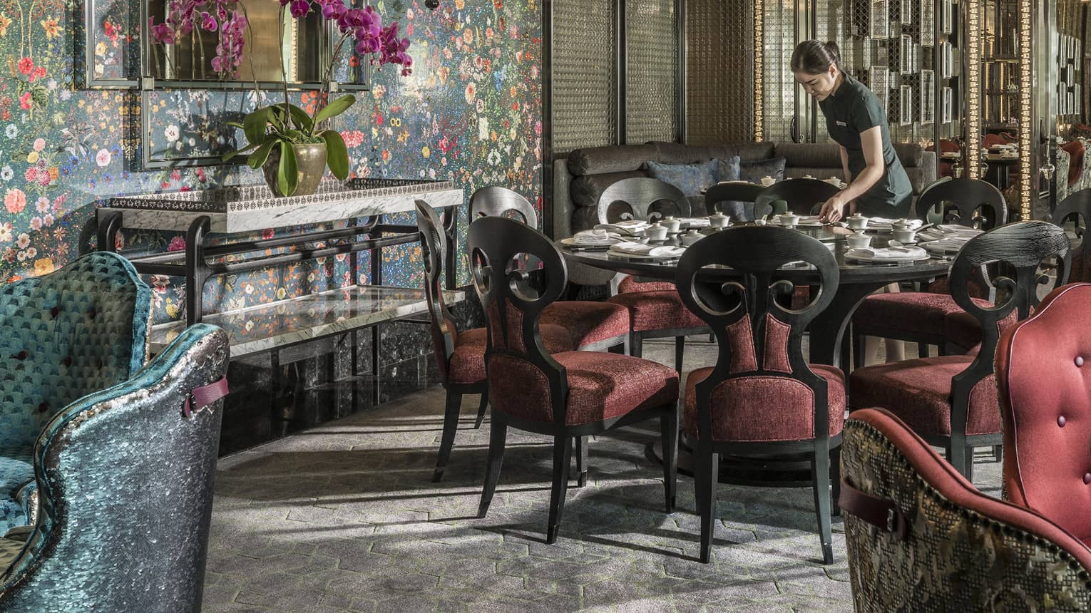 Hotel staff sets table with elegant red velvet chairs under tall mirror, floral wallpaper 