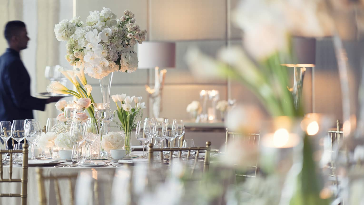 A four seasons staff member sets the tables of a wedding reception with white tall florals, clear glassware, white plates and silver tableware