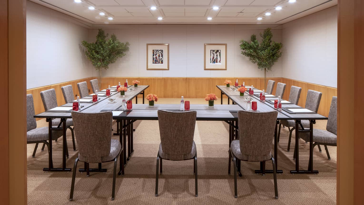 Multiple long, rectangular boardroom tables are placed together to form a "U" shape facing blank wall and are set with a black folder, white paper, pen and clear bottles filled with a red soda. 