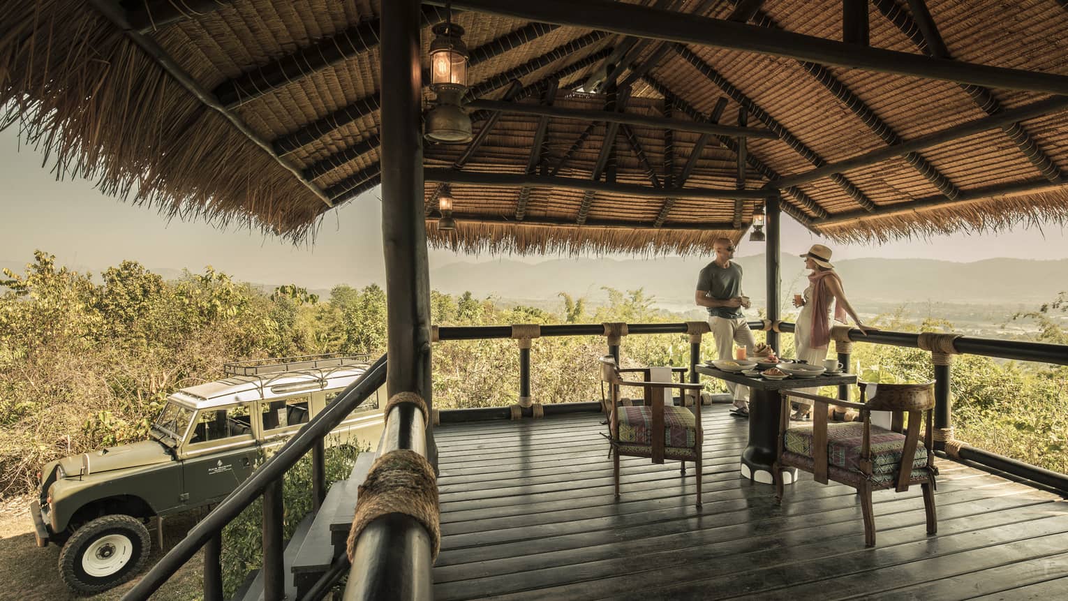 A couple sitting on a wooden deck overlooking green brush, Land Rover sitting nearby 