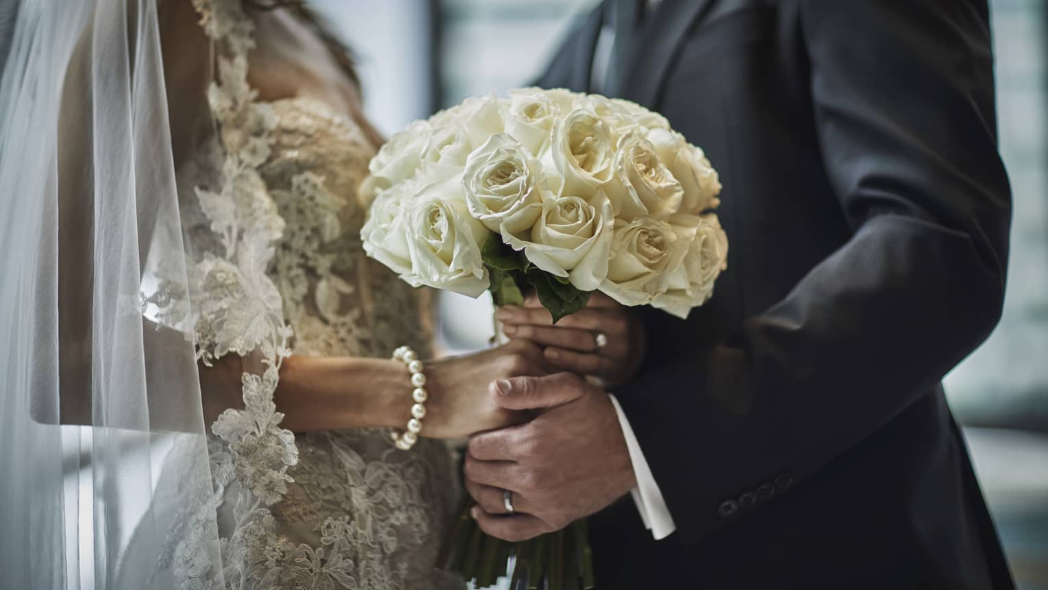 A close up of a bride in a lace gown and a groom in a gray suit hold the bride's bouquet of white roses together.