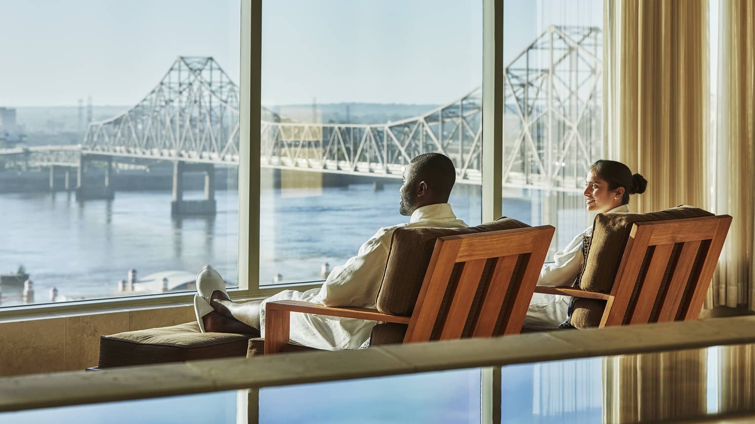 Couple lounge in chairs by large windows overlooking water, St. Louis bridge